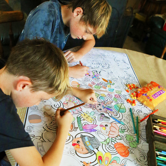 Two children coloring a large thanksgiving sheet of paper with markers on a table.