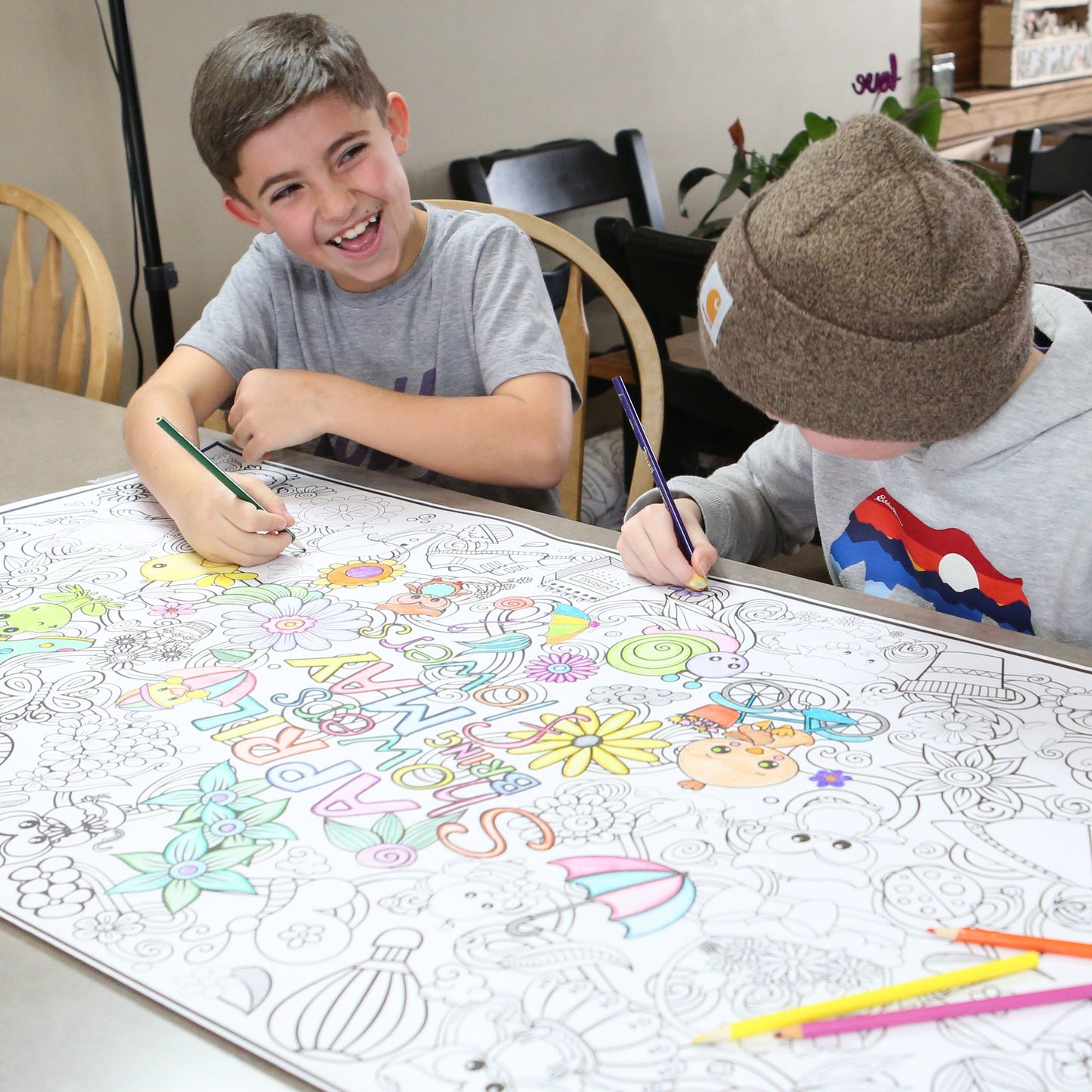 Two children coloring on a large sheet of paper with markers at a dining table.