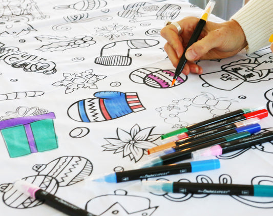 A Christmas-themed coloring tablecloth laid out on a table with various Christmas motifs such as trees and ornaments, accompanied by a bowl of coloring pens and decorative items.