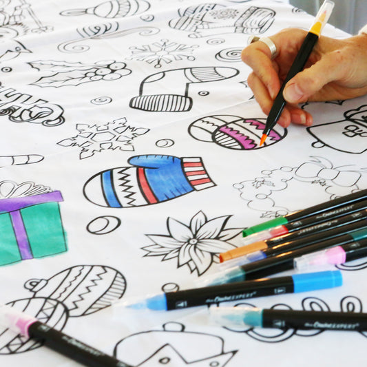 A Christmas-themed coloring tablecloth laid out on a table with various Christmas motifs such as trees and ornaments, accompanied by a hand coloring with markers.