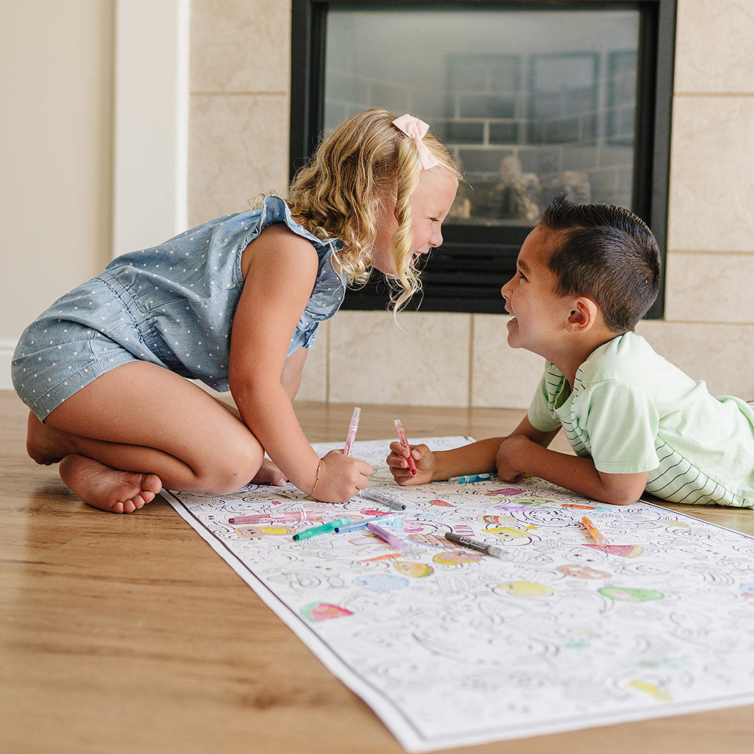 Two children sitting on the floor in front of a fireplace, engaged in an activity with a fun food coloring poster and crayons.