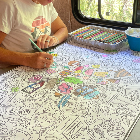 Person coloring a large sheet of paper with crayons on a table inside an RV.