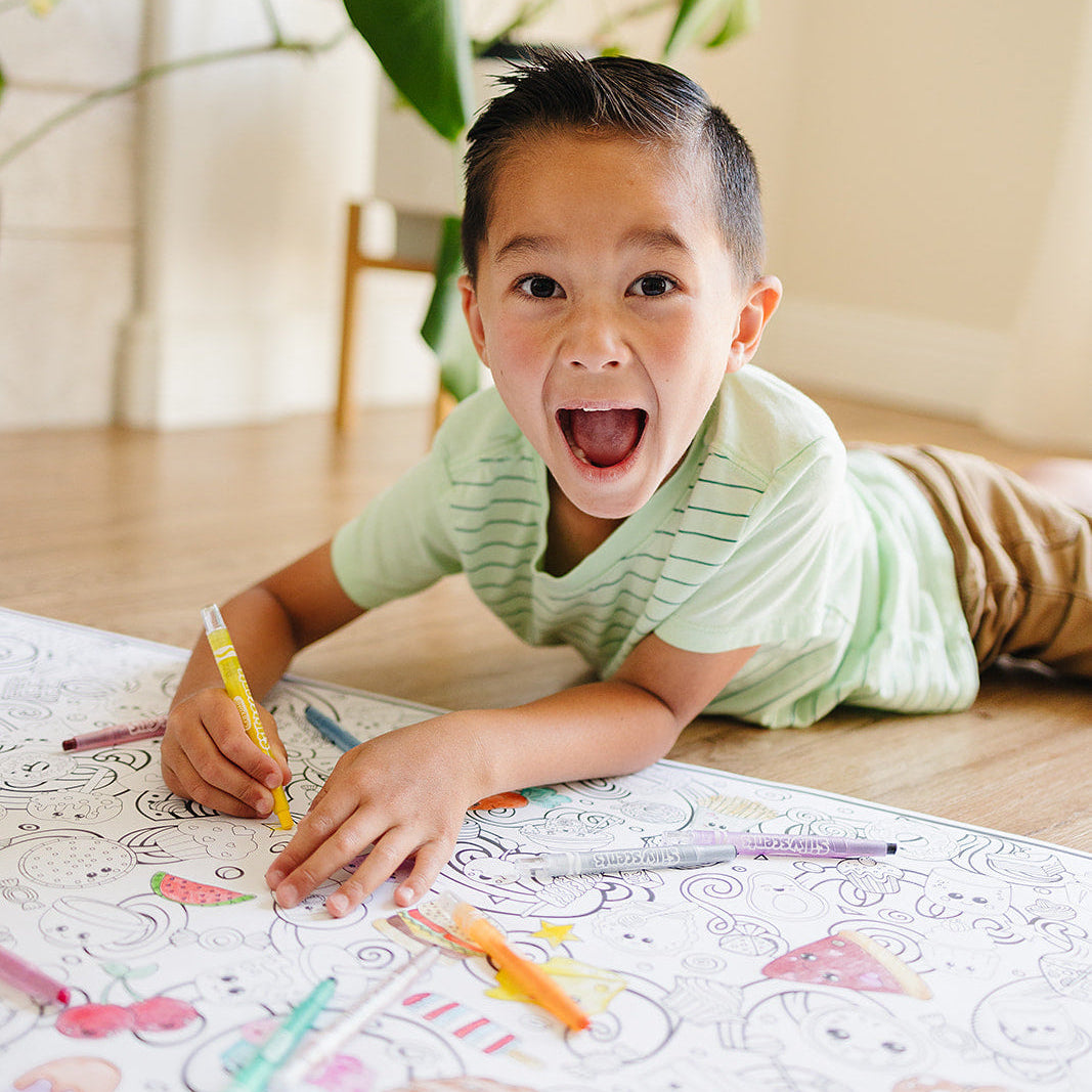 Child lying on the floor with coloring book and crayons, surrounded by green plants.