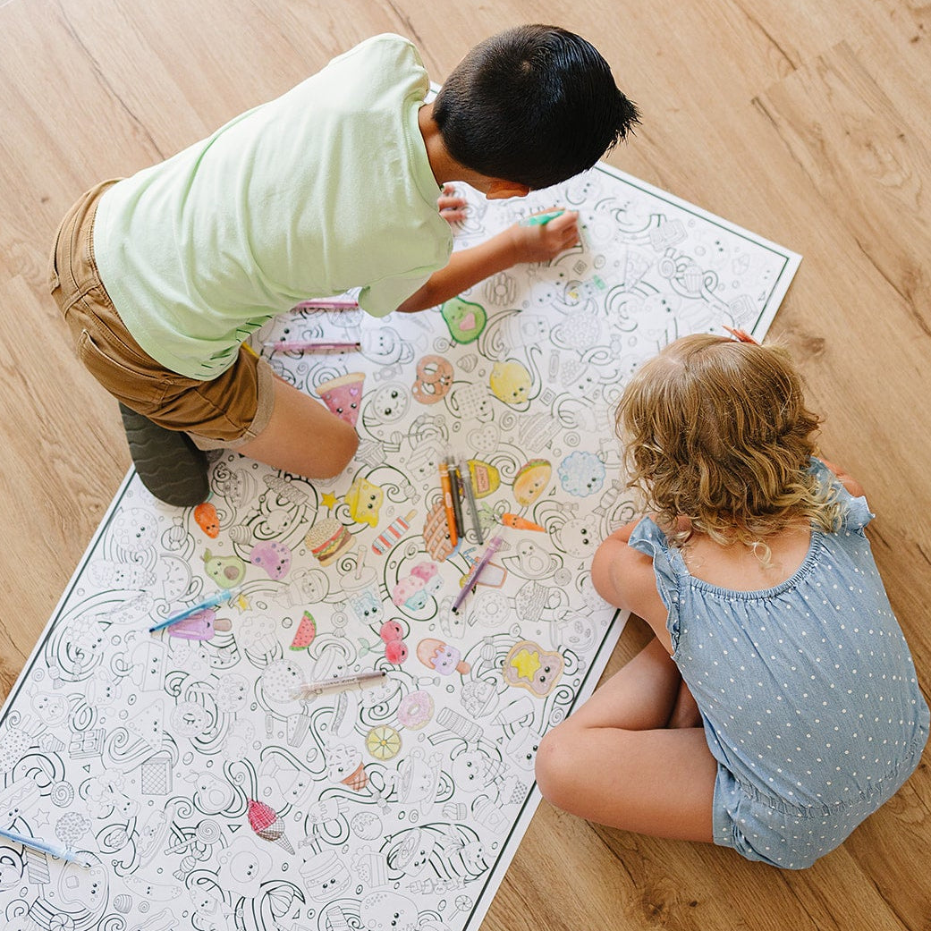 Two children coloring on a large coloring sheet on a wooden floor.