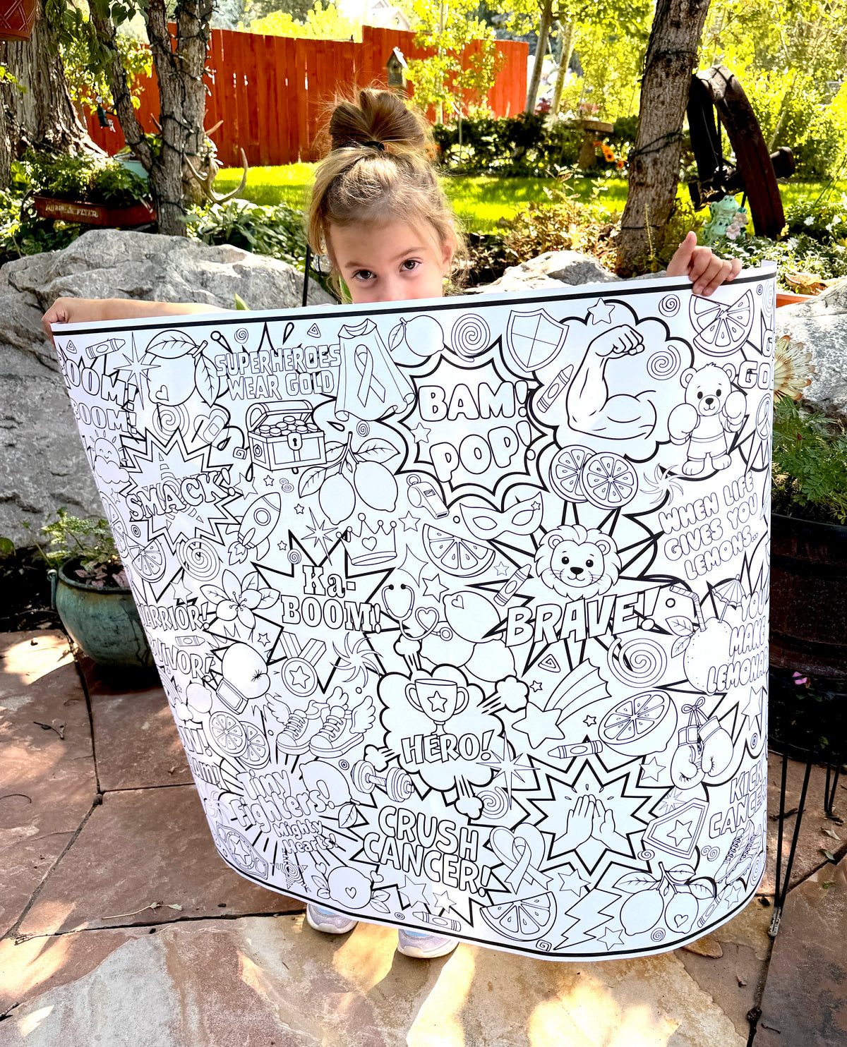 Kick Kids' Cancer Coloring Poster held by a little girl. A dense, colorful doodle-style graphic filled with motivational words like BOOM, BAM!, SMACK, WARRIOR, HERO, BRAVE, and YOU MAKE LEMONUS, plus illustrated elements such as trophies, stars, hearts, lemons, rainbows, and sports icons on a white background.