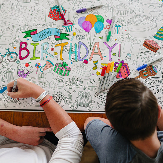 Two people coloring a large sheet of paper with birthday-themed doodles.