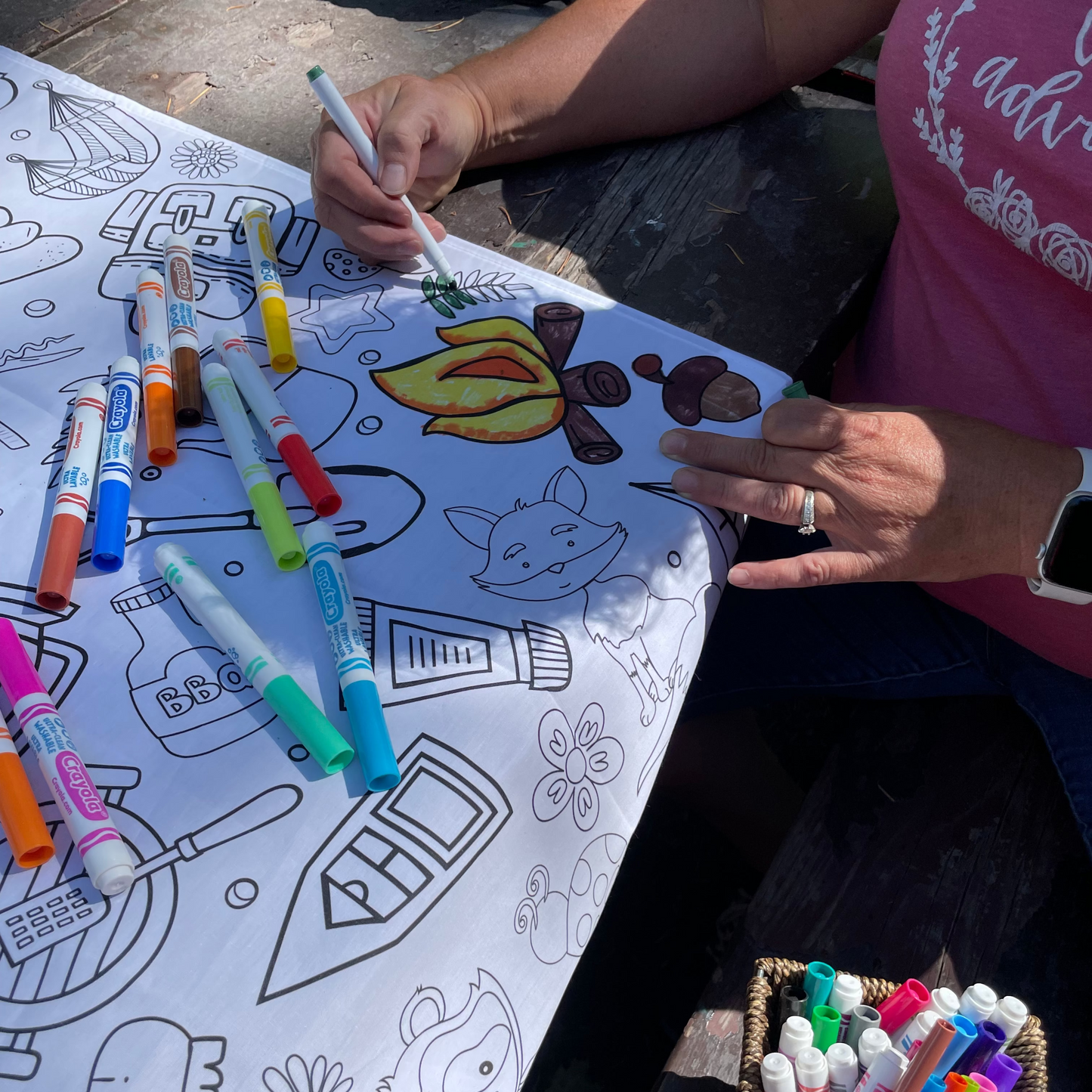 A lady coloring on an Artistic Chaos Ink Camping Fabric Reusable coloring tablecloth with a green marker. Tablecloth has a campfire image and animals
