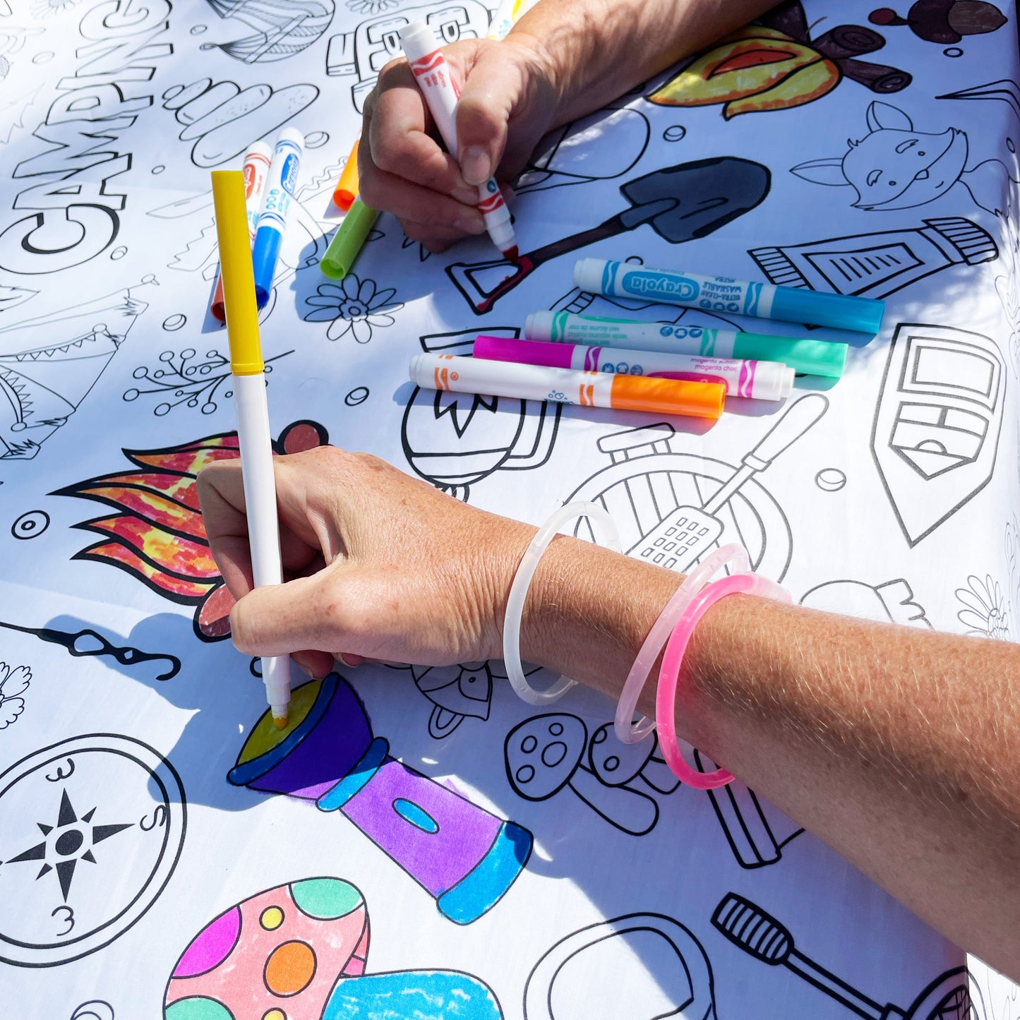 Hands holding markers while coloring an Artistic Chaos Ink Camping Coloring Tablecloth. One hand is holding a yellow marker and coloring a flashlight. The other hand is holding a red marker coloring a shovel.