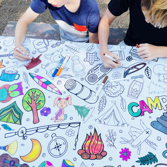 Two individuals coloring on a white fabric coloring tablecloth with various camping-themed illustrations such as trees, mountains, and camping gear and animals. They are sitting outside on a camp table.