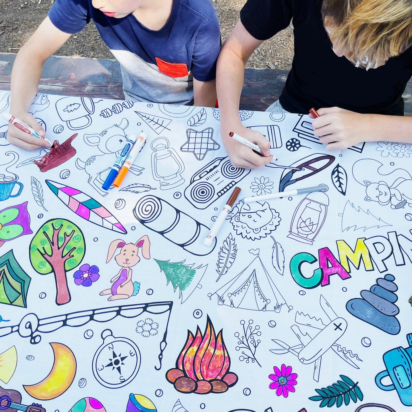 2 boys coloring on a fabric coloring tablecloth with markers. The fabric coloring cloth has images of camping gear, animals and nature.