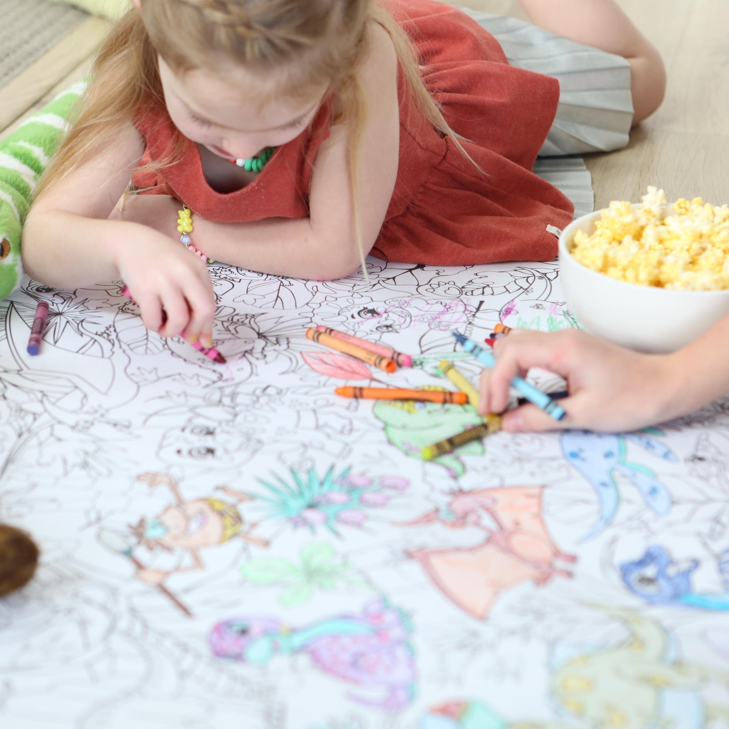 Child coloring on a large dinosaur themed coloring poster with crayons, surrounded by toys and snacks.