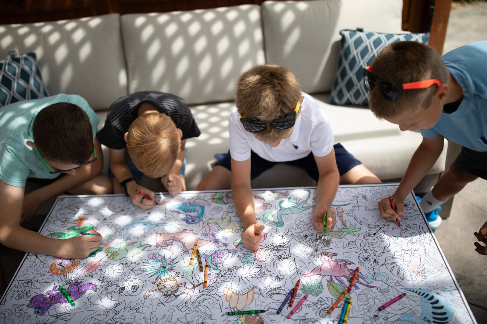 Four children gathered around a table outdoors, coloring a large illustrated sheet with crayons. The scene is set in a real environment with a sofa in the background, making the subject type Lifestyle.