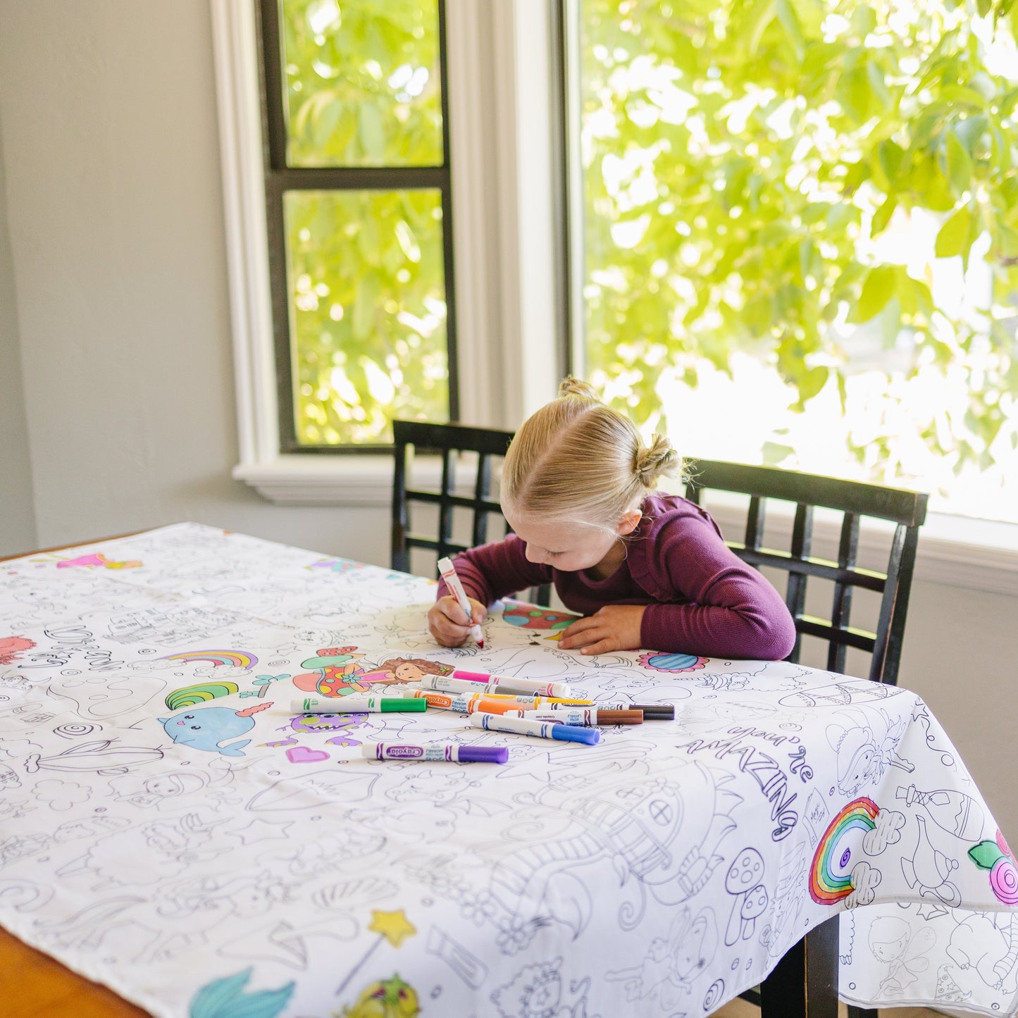 Little girl sitting at a table coloring with markers on a fabric coloring tablecloths with whimsical characters and magical creatures. 