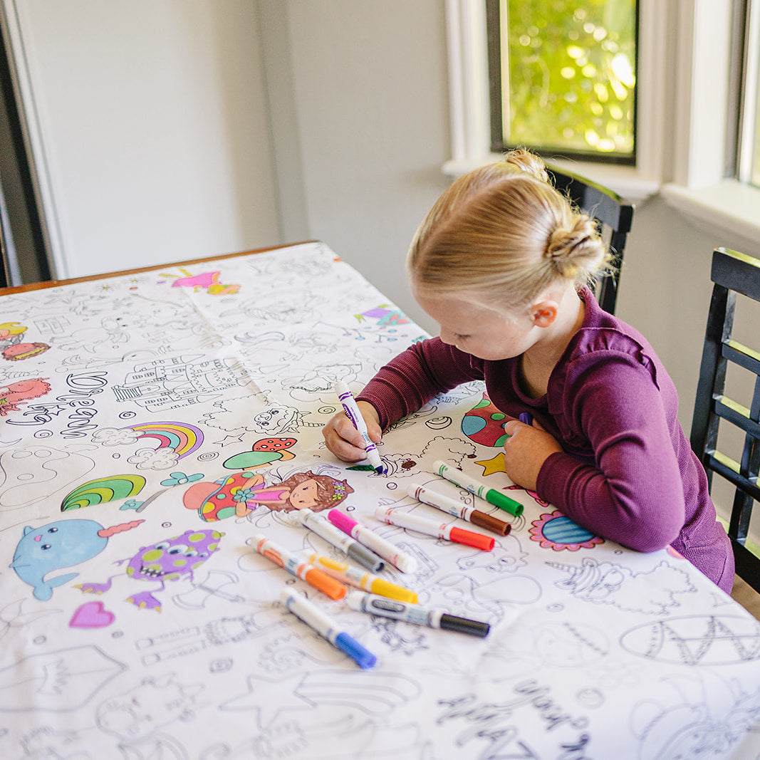 Child with pigtails coloring on a table with a fabric coloring tablecloth on it and  markers in a bright room