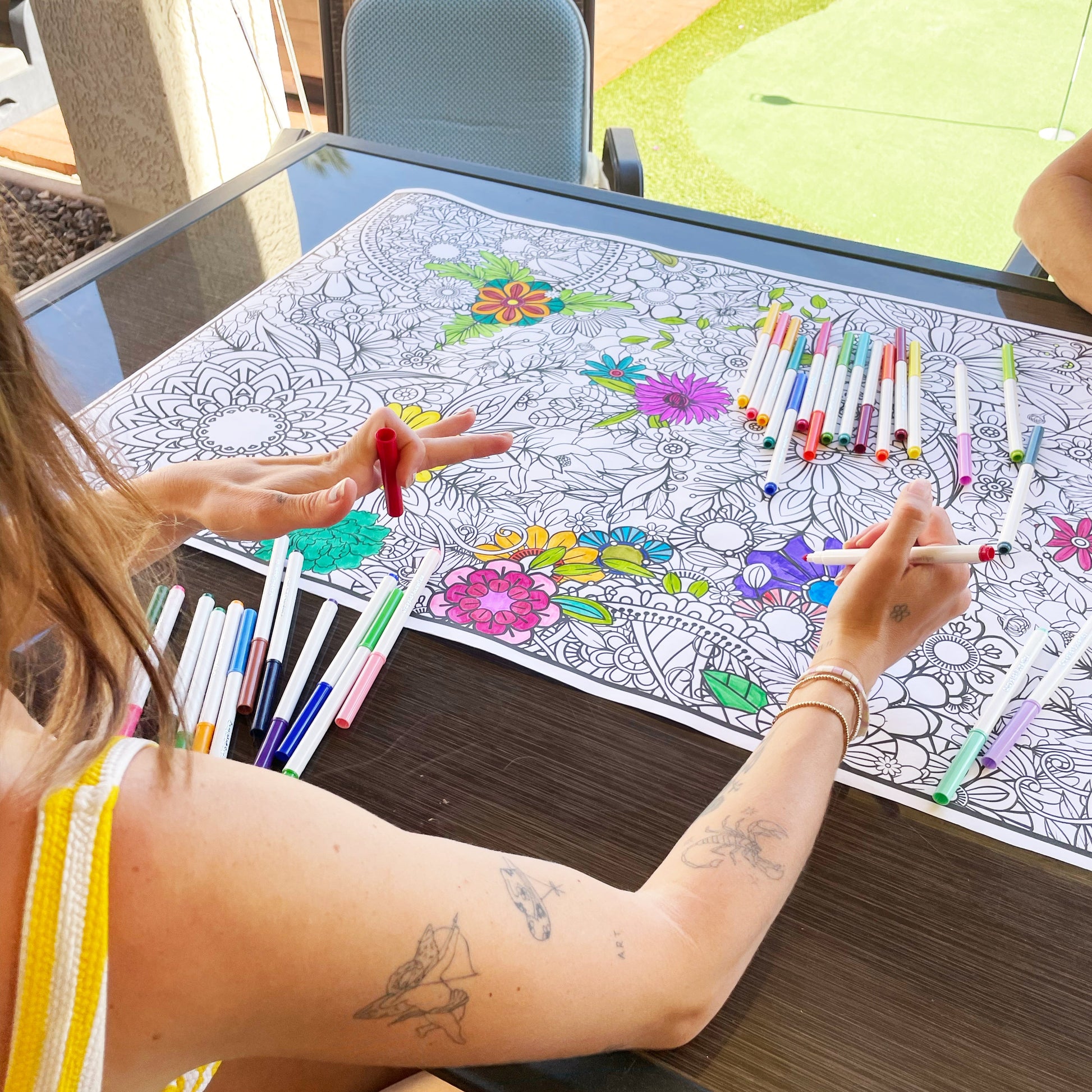 Person coloring a floral design on paper with markers at a table.