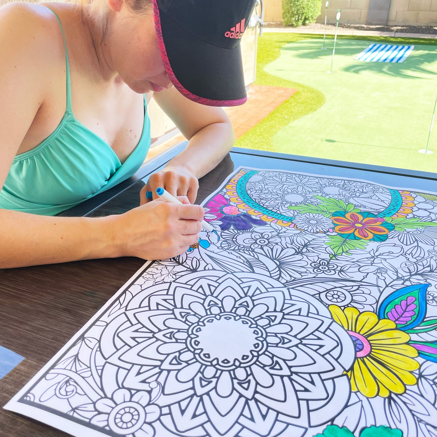 Person coloring a mandala on an outdoor table with a pool in the background