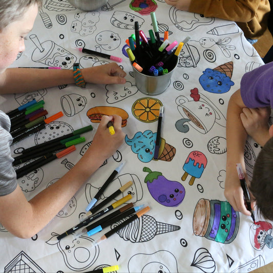 two boys coloring a food tablecloth with washable markers. The food has funny faces
