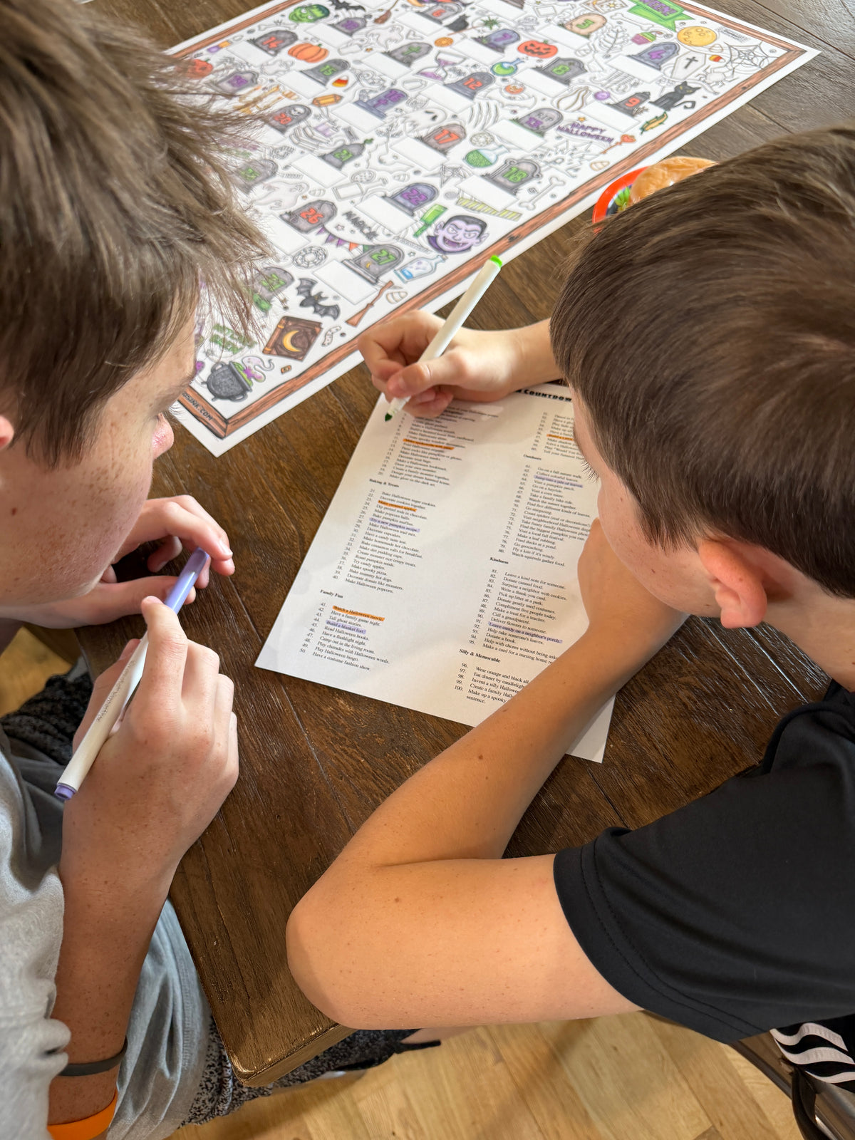Two boys sitting at a wooden table, writing on a checklist with pens. A large illustrated activity board is spread out on the table in the background.
A black and white Halloween advent calendar COLORING POSTER titled 'Creepin' to Halloween!' with 25 numbered tombstone boxes and many spooky doodles throughout.  Halloween Countdown coloring poster from Artistic Chaos Ink.