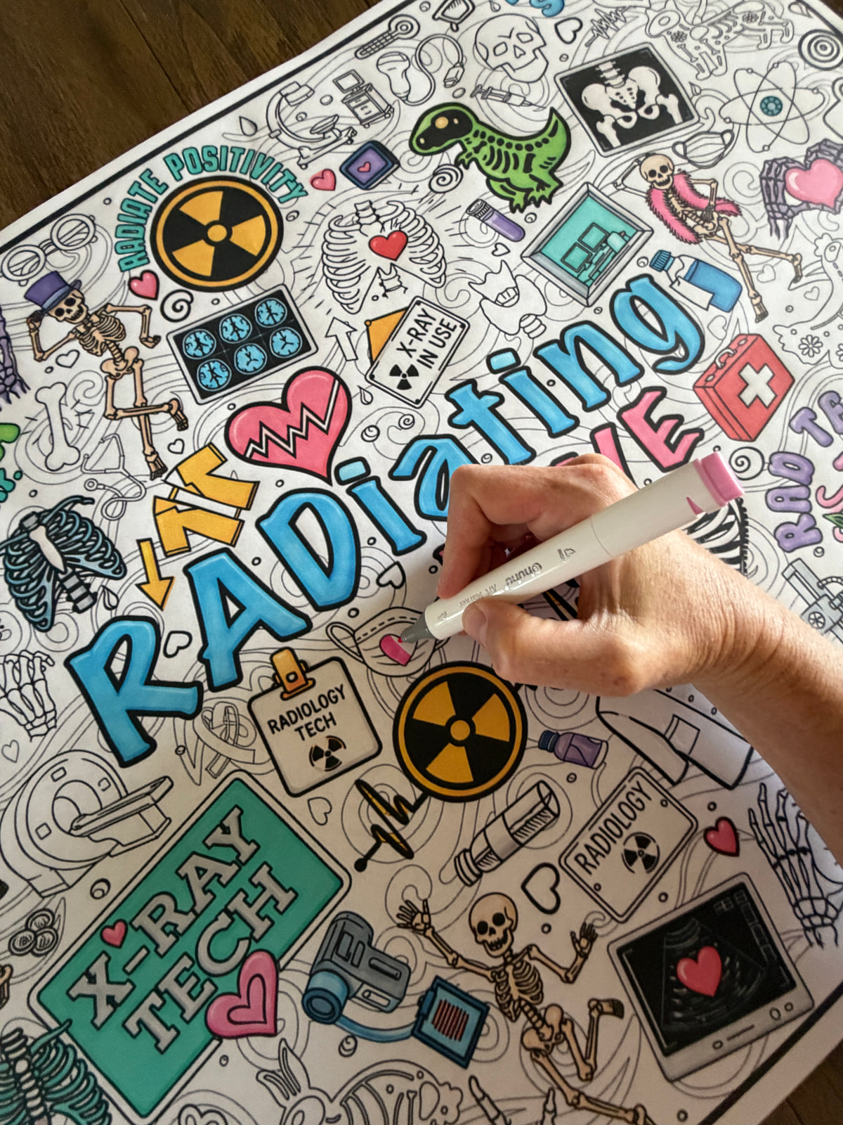 A hand holding a pink marker is coloring a detailed black-and-white poster about radiology and X-rays, with colorful illustrations and the word 'RADIATING LOVE' in large blue letters. The scene is photographed from above on a wooden surface. Radiating Love coloring poster from Artistic Chaos Ink.