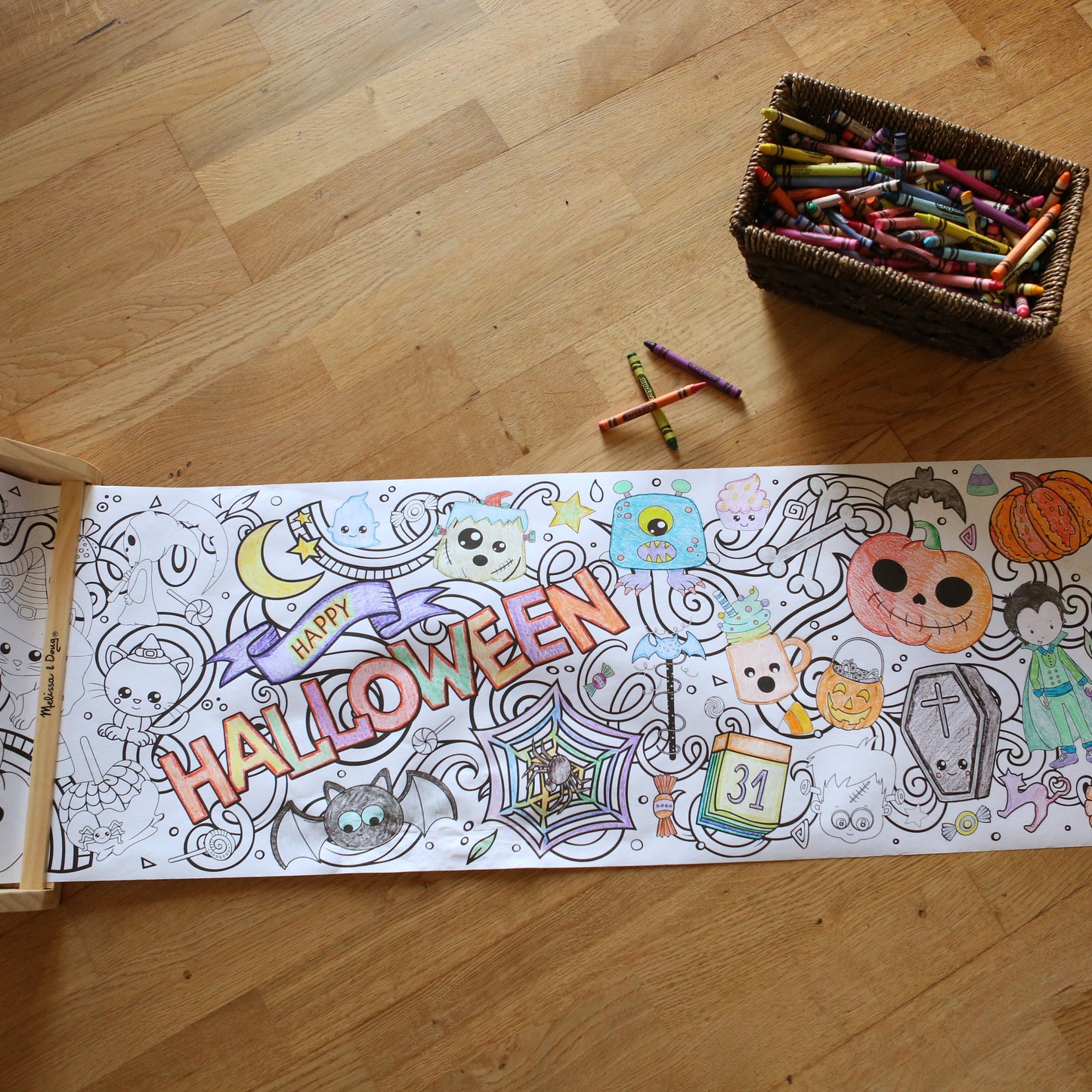 Long Coloring sheet with Halloween-themed illustrations on a wooden floor with crayons.