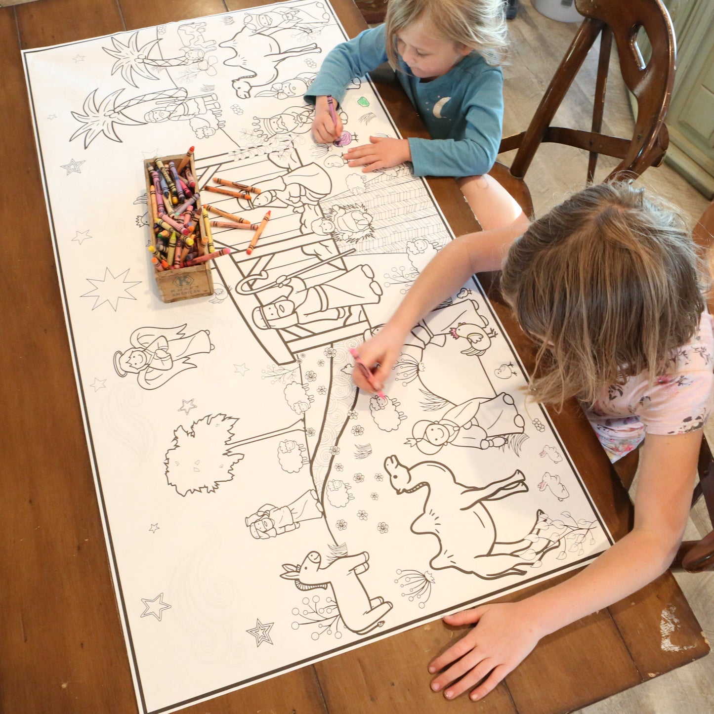 Two children coloring on a nativity coloring large sheet of paper with crayons at a wooden table.