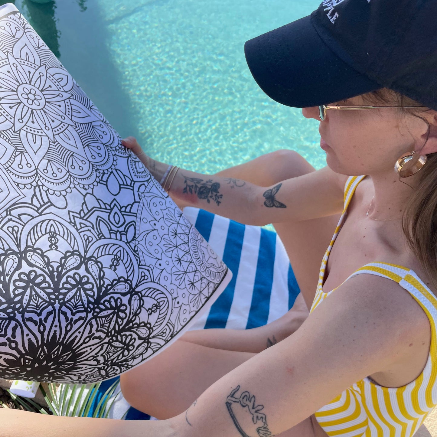 Person sitting by a pool holding a coloring book with a mandala design.