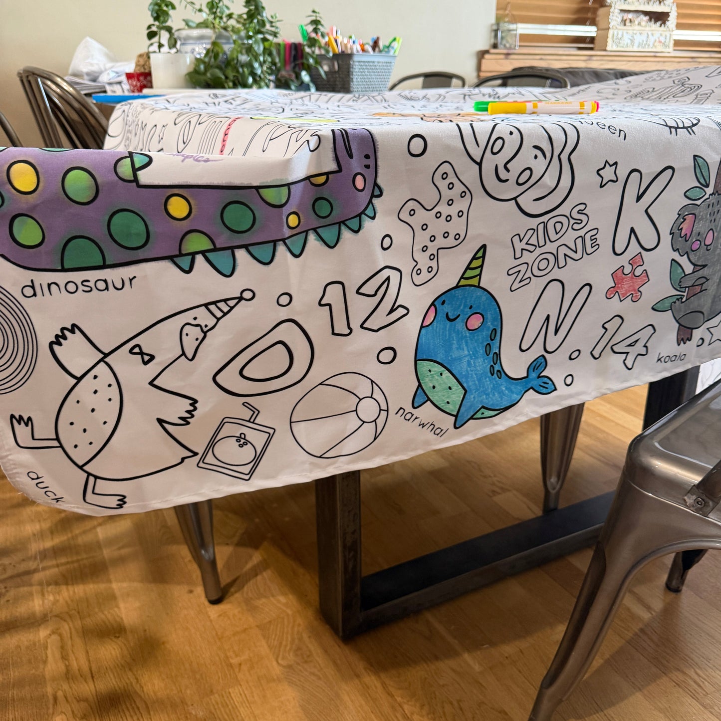 Children's fabric coloring tablecloth with colorful dinosaur design on a wooden table.