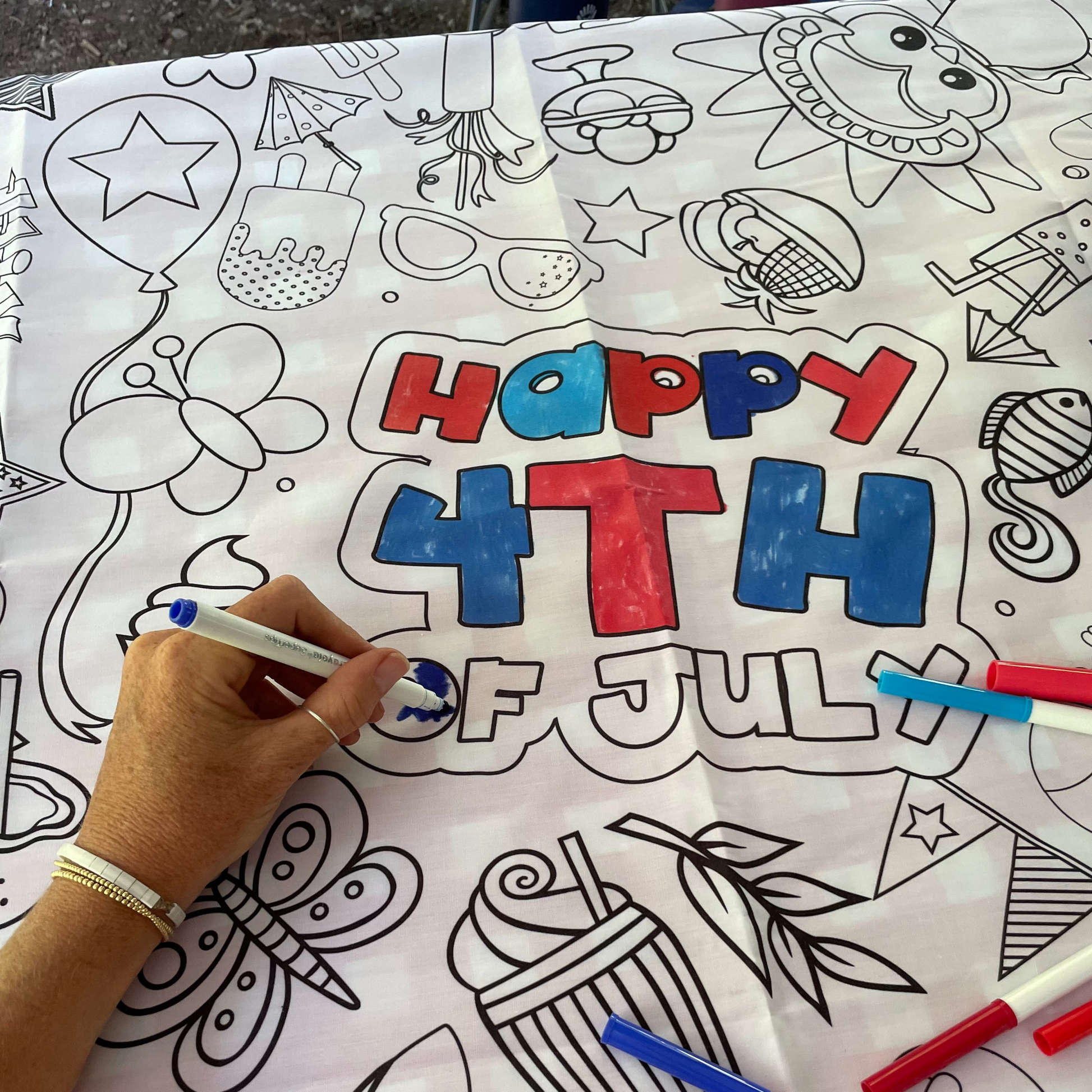 Coloring fabric tablecloth with 'Happy 4th of July' text and patriotic illustrations, being colored by a hand with red and blue markers.