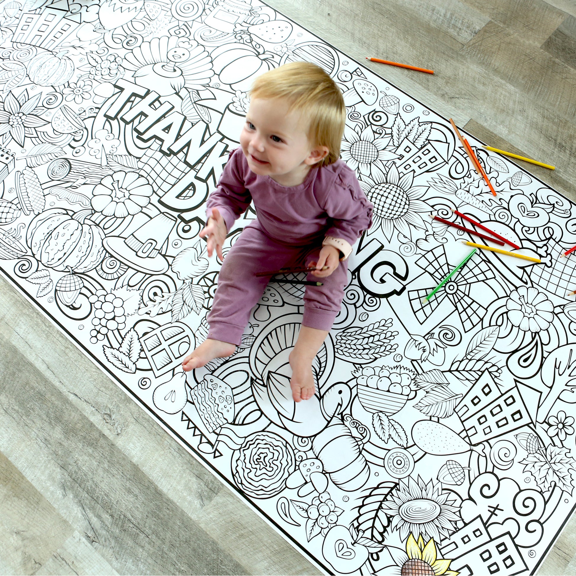 large coloring banner on the floor with small baby sitting on it to show size. Coloring banner has fall and thanksgiving graphics on it. 