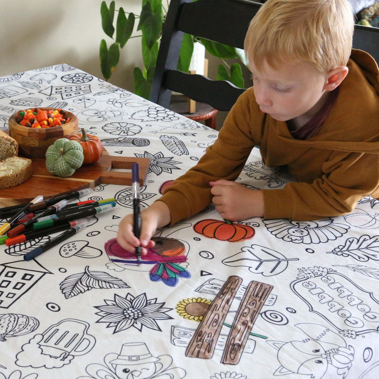 A child is coloring on a white fabric tablecloth decorated with Thanksgiving-themed graphics, including turkeys, pumpkins, and leaves. The tablecloth is laid out on a table with a variety of coloring markers and crayons.