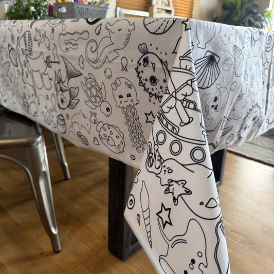 White under the sea fabric coloring tablecloth with black fish and shell designs laid on a table to be colored in a room with plants and a window.