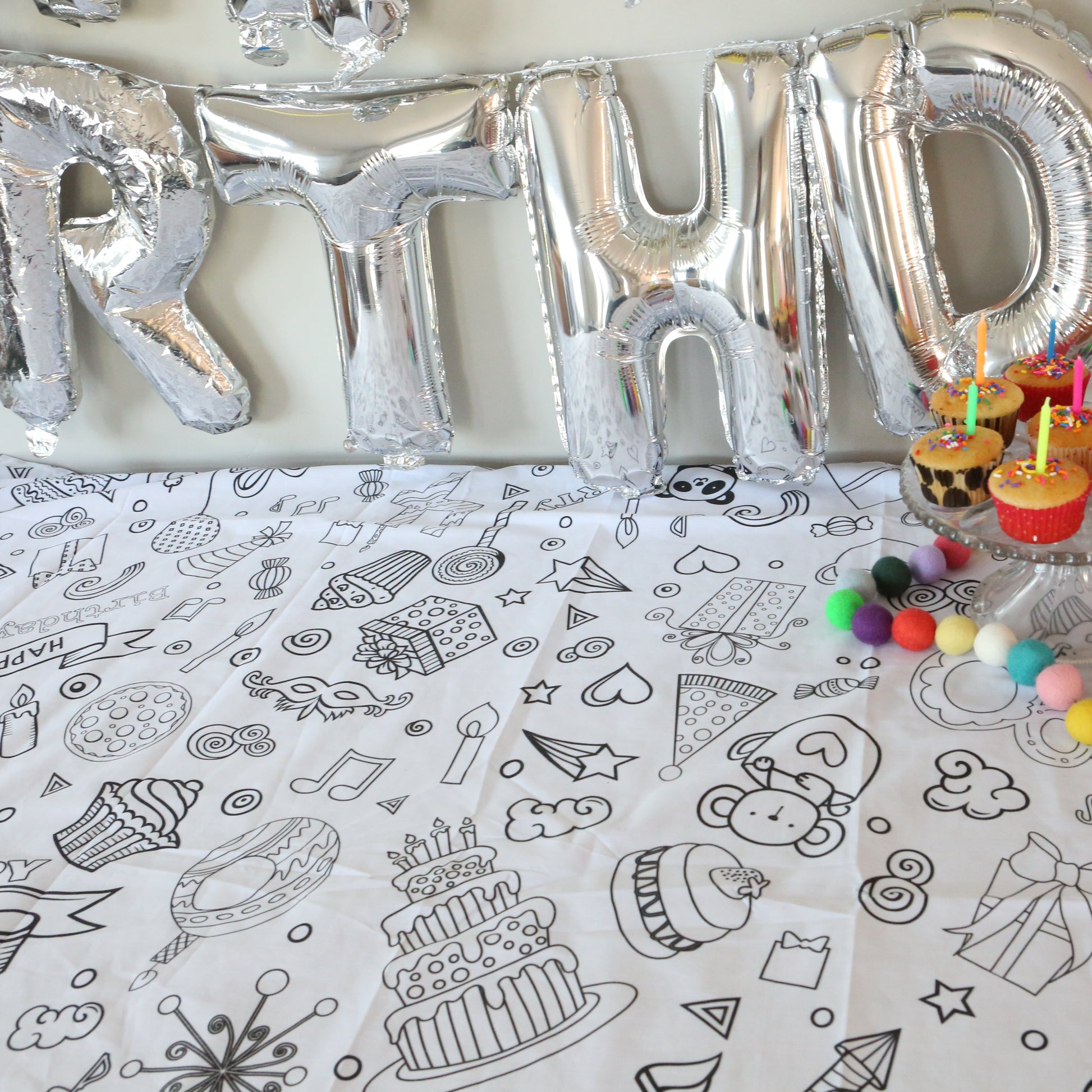 Silver 'BIRTHDAY' balloons and small birthday cakes with candles on an Artistic Chaos Ink coloring tablecloth with black and white designs.