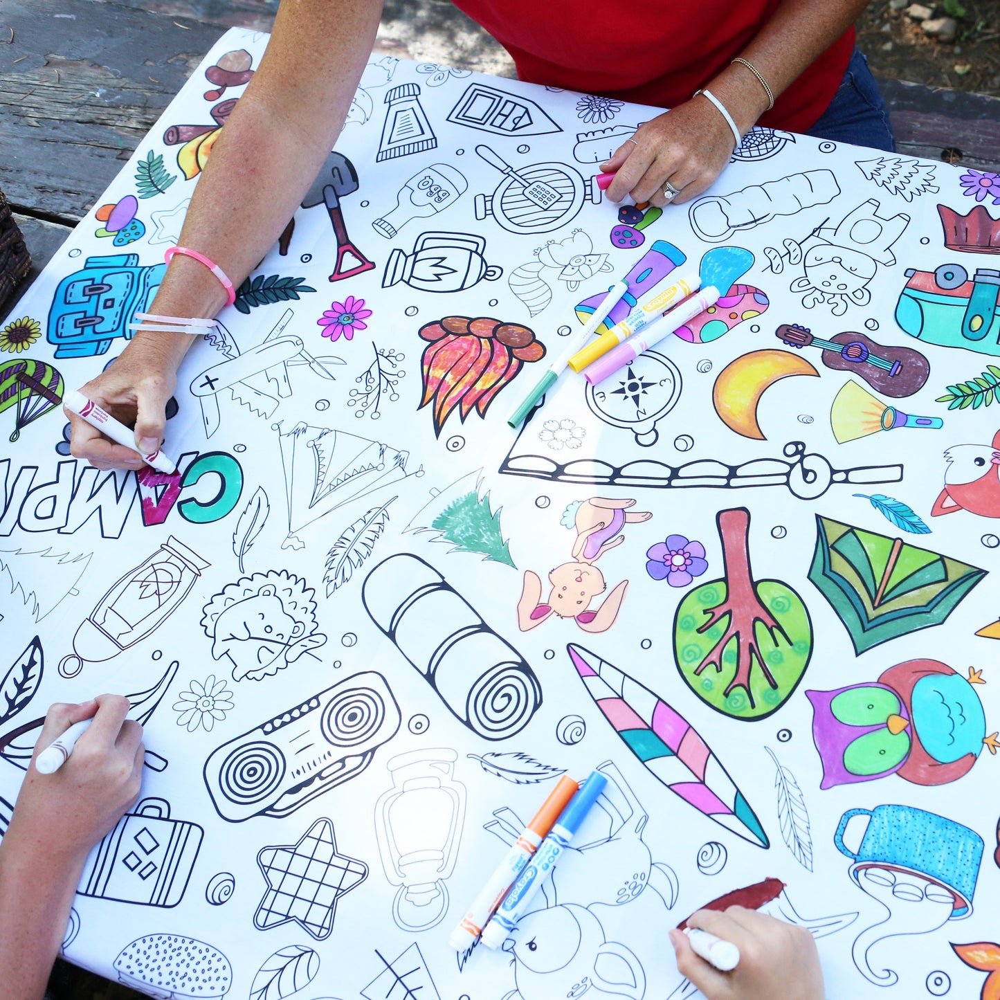 People coloring on a large tablecloth with various camping designs and objects.