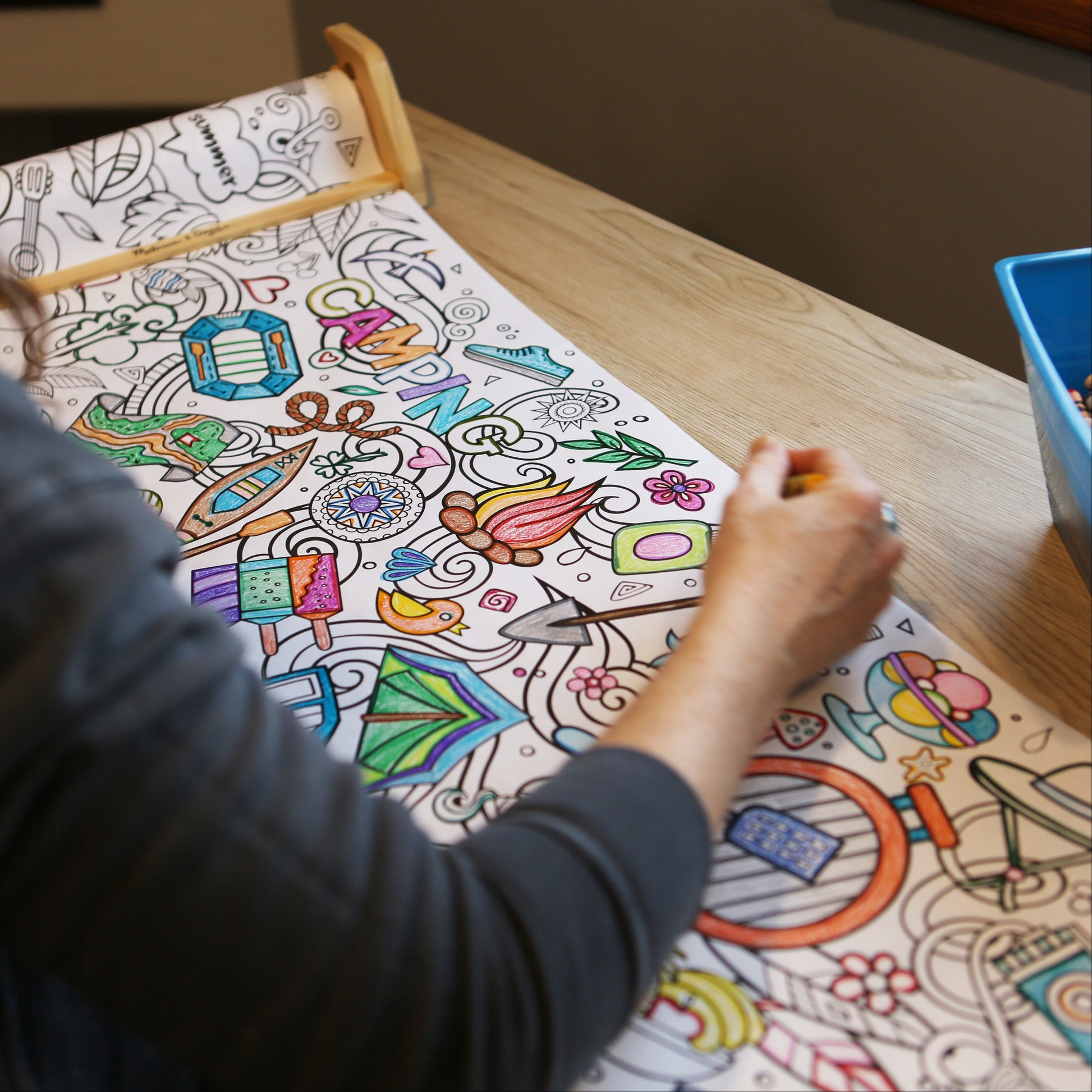 Person coloring a detailed coloring summer coloring roll with crayons on a wooden table.