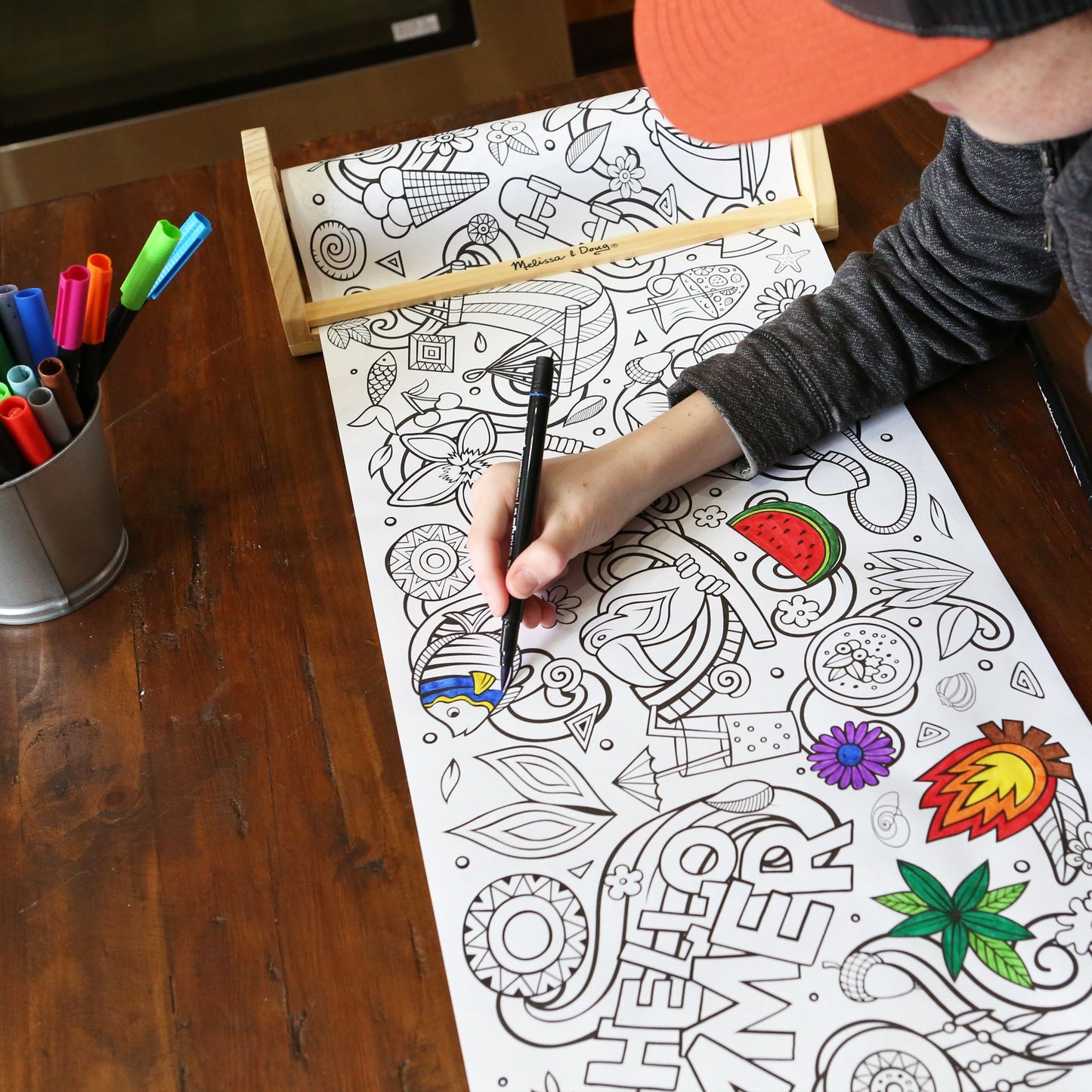 Boy with hat coloring with a marker on a summer time coloring sheet. Watermelon, fish plants and popsicles are among the graphics.