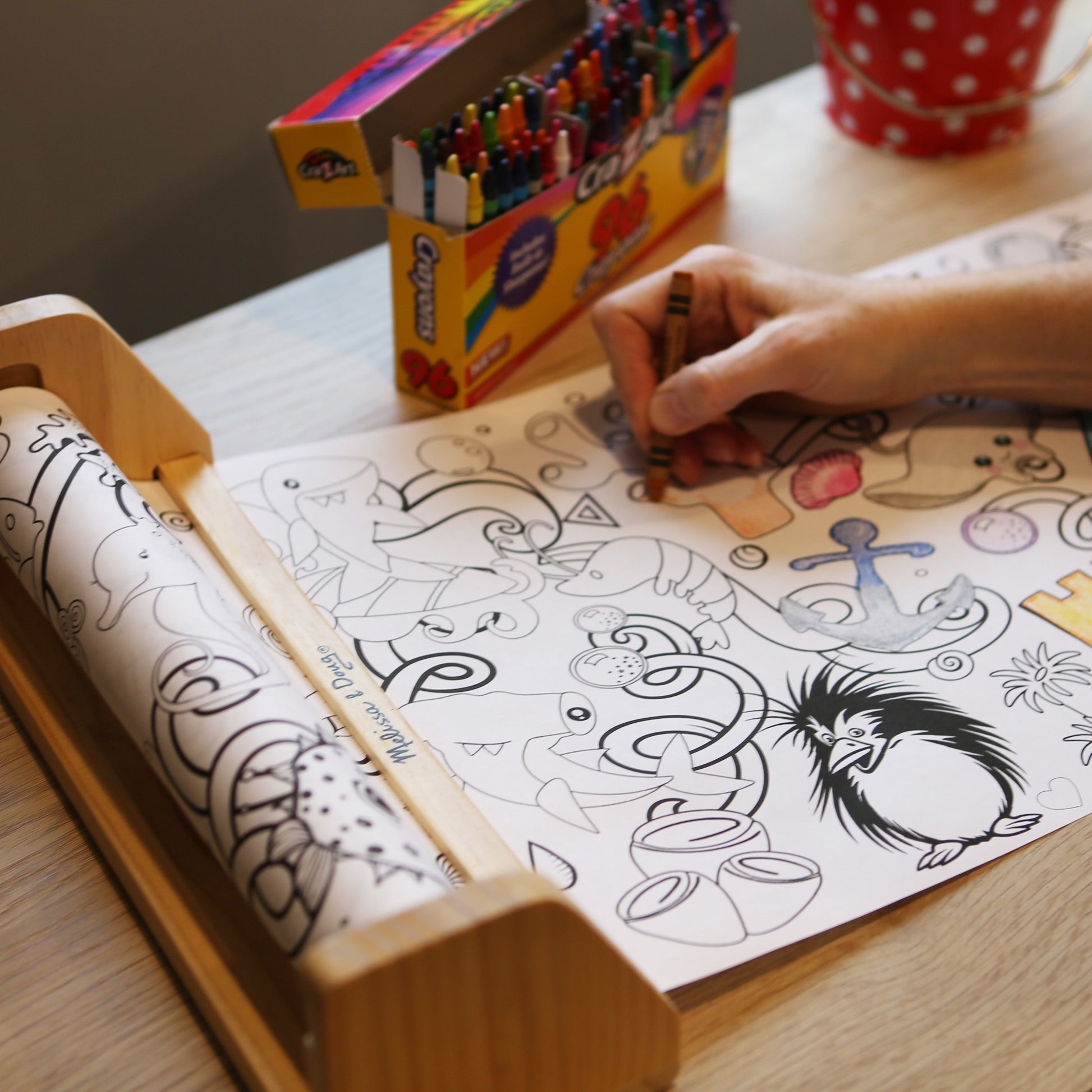 Person coloring a picture on a table with a box of crayons and a mug in the background.