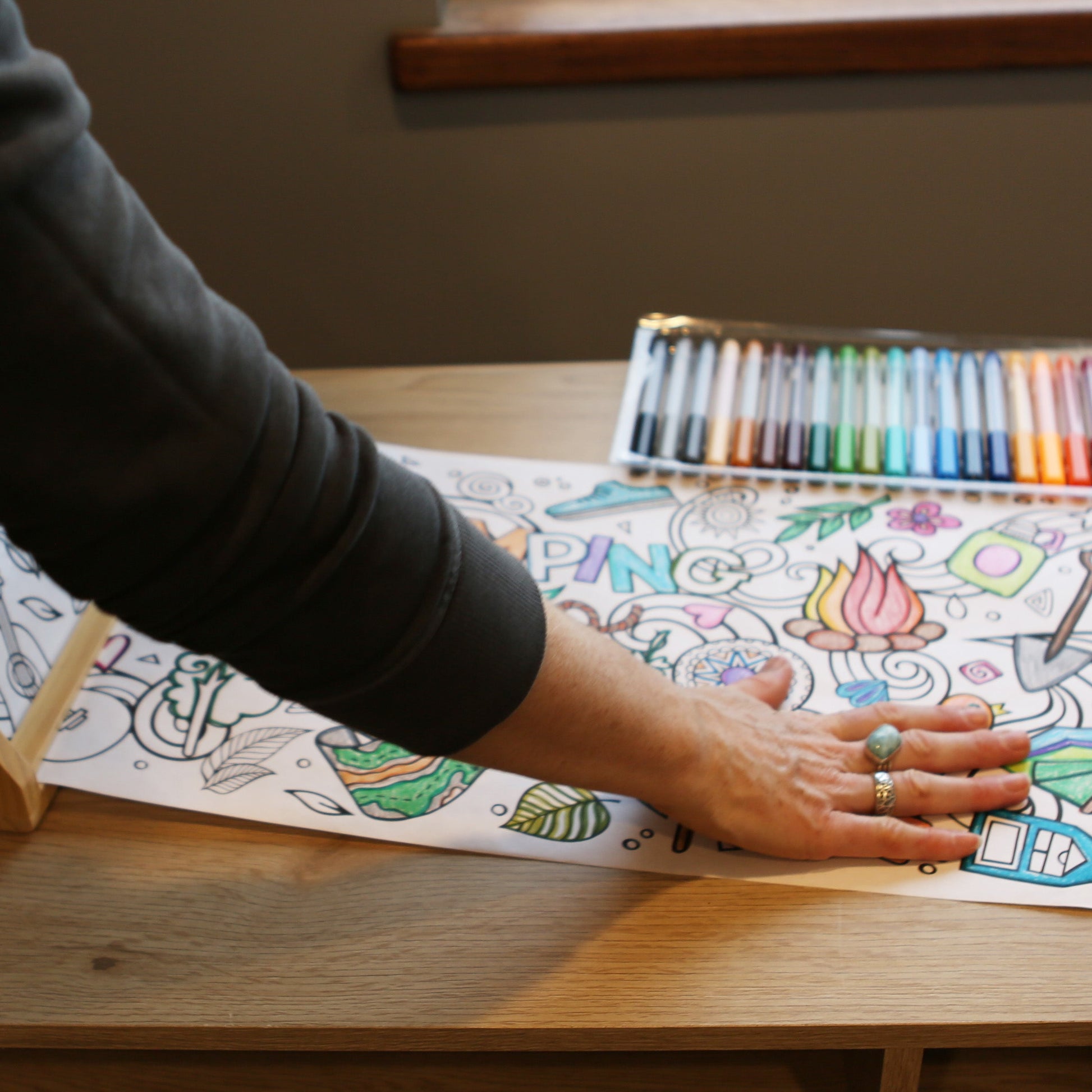 Person coloring a detailed design on a large sheet of paper with colored pencils on a wooden table.
