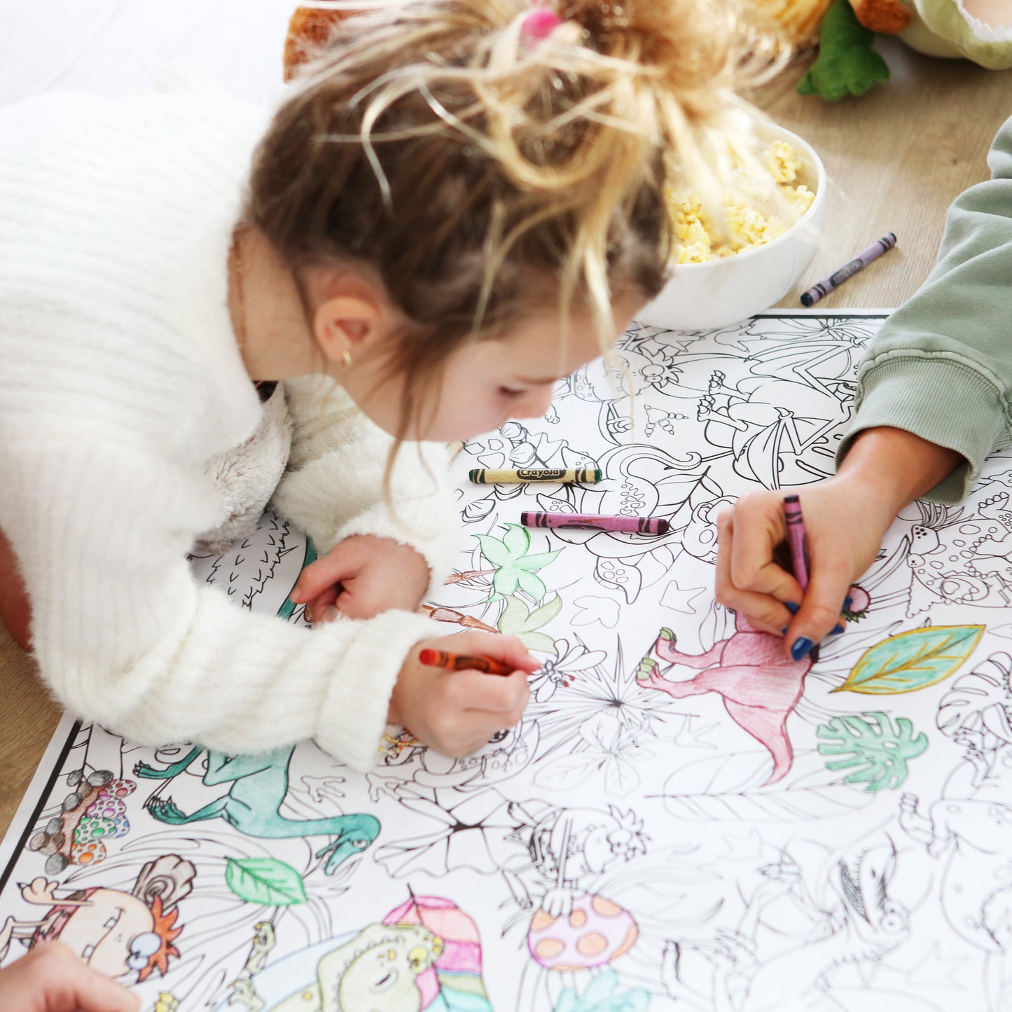 Two children coloring on large sheets of paper with markers on a wooden floor.