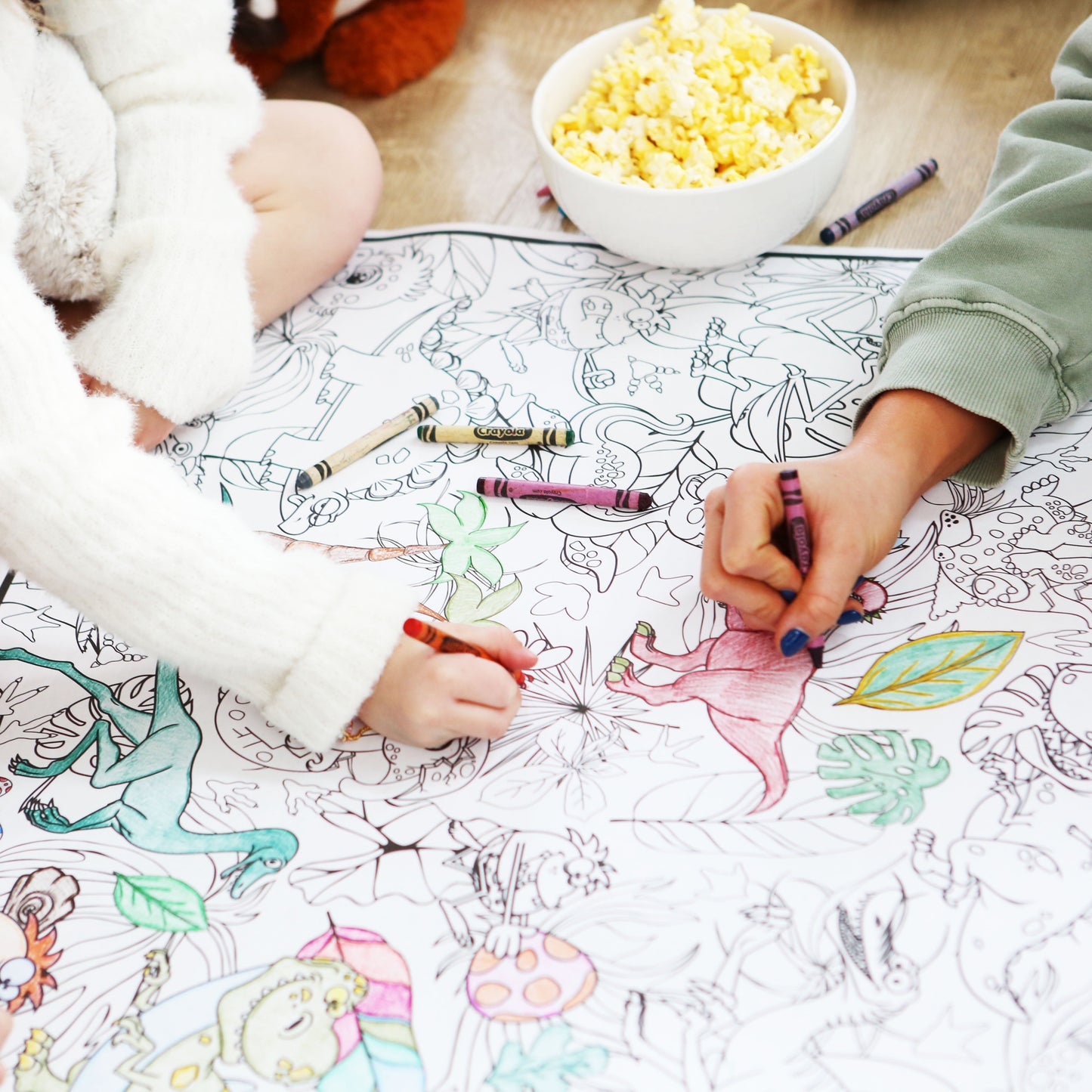 Children coloring on a large sheet of paper printed with dinosaurs with crayons, sitting around a bowl of popcorn