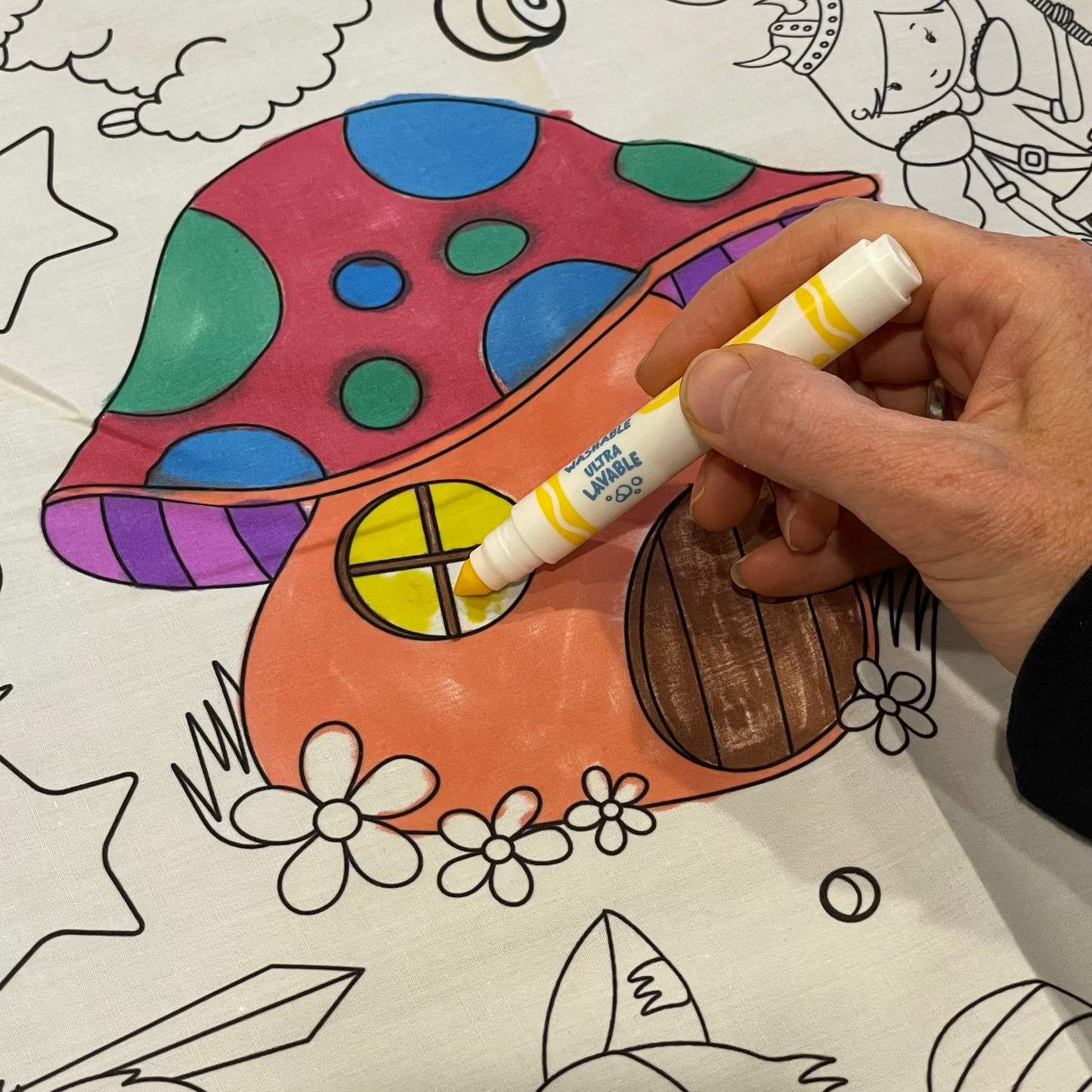 Artistic Chaos Ink Coloring fabric tablecloth with a mushroom house drawing and a hand holding a marker.