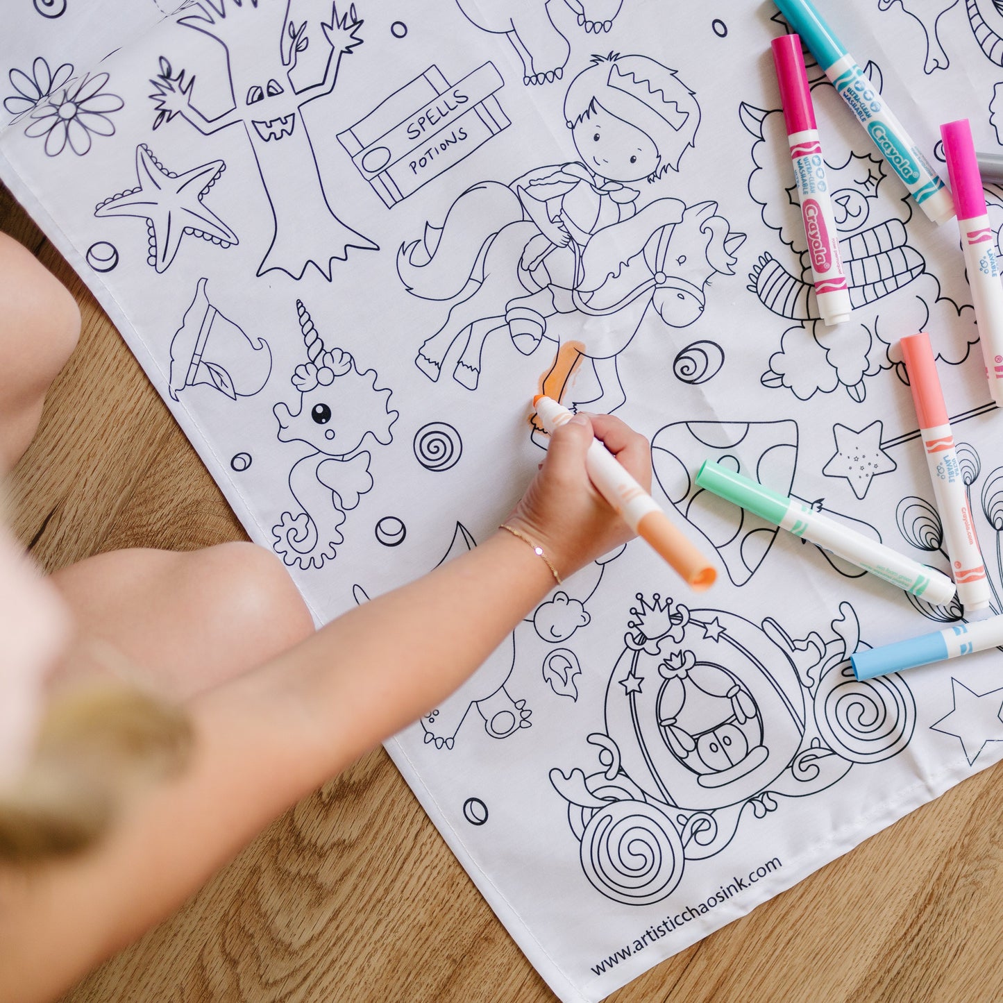 Child coloring a unicorn-themed coloring tablecloth with markers on a wooden floor.