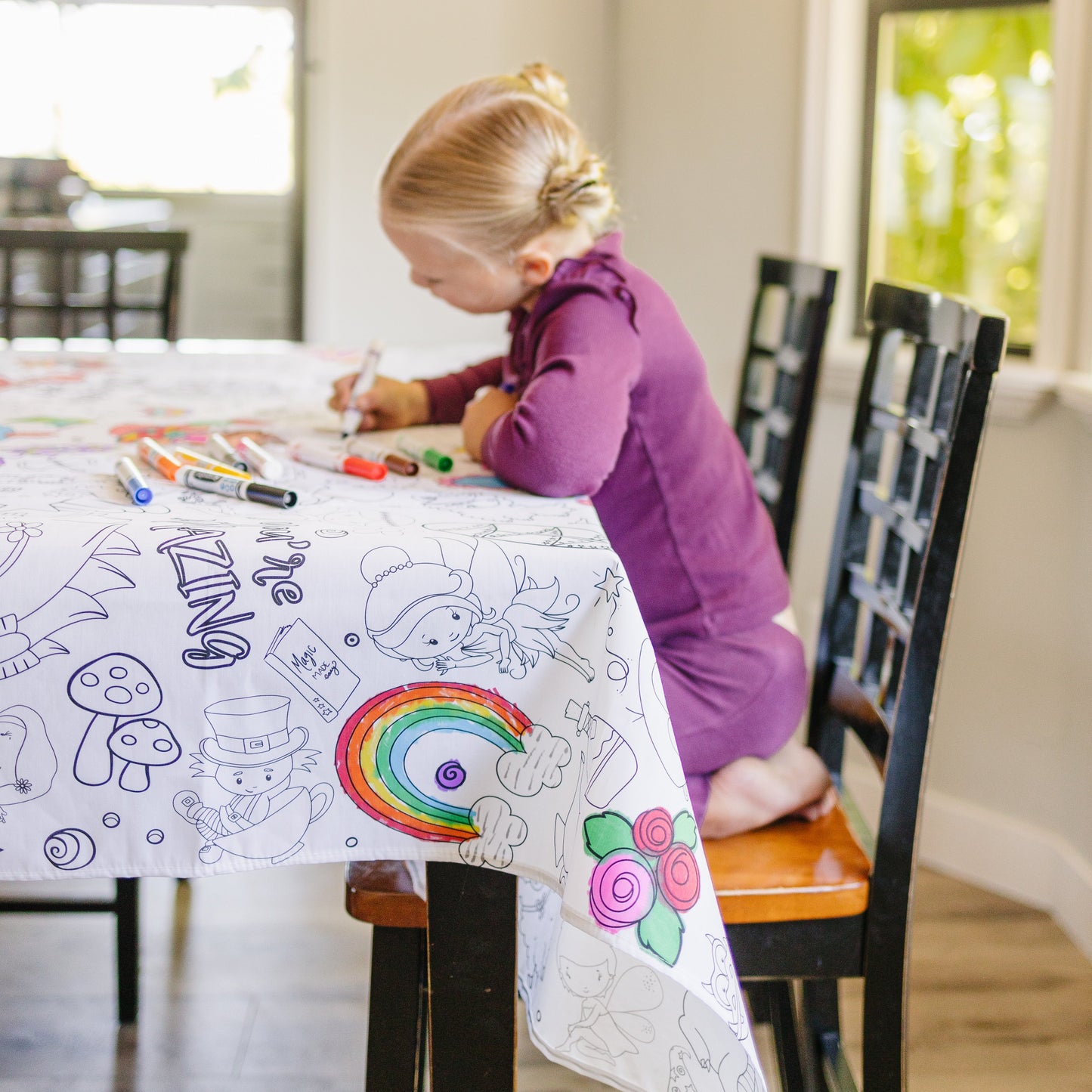 A child coloring on a white and black fabric tablecloth with various colorful drawings including rainbow, fairy, flowers, mushroom, placed on a table with chairs around it.
