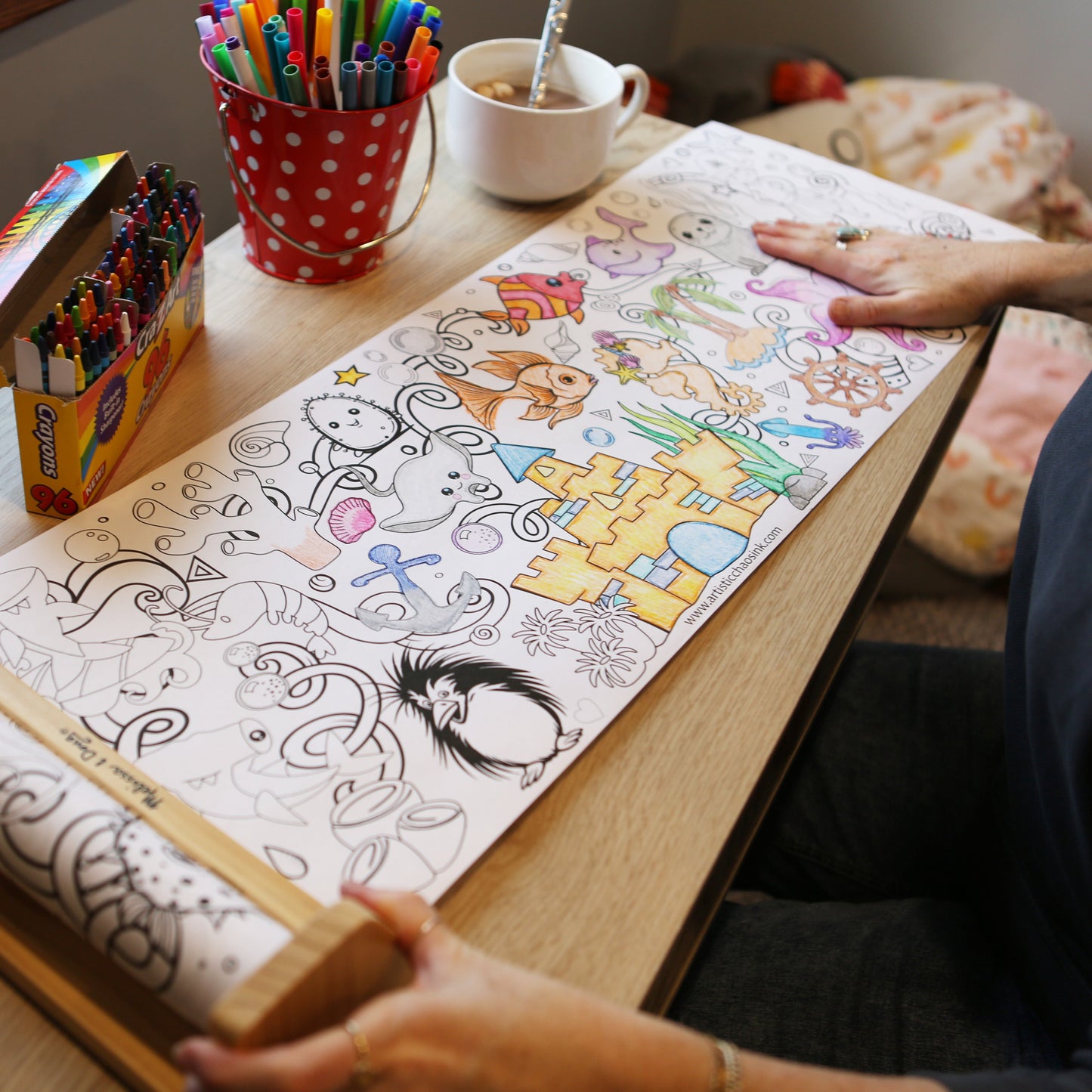 Person coloring a large sheet of paper with markers and a cup of tea on a wooden table. Coloring roll is by Artistic Chaos Ink