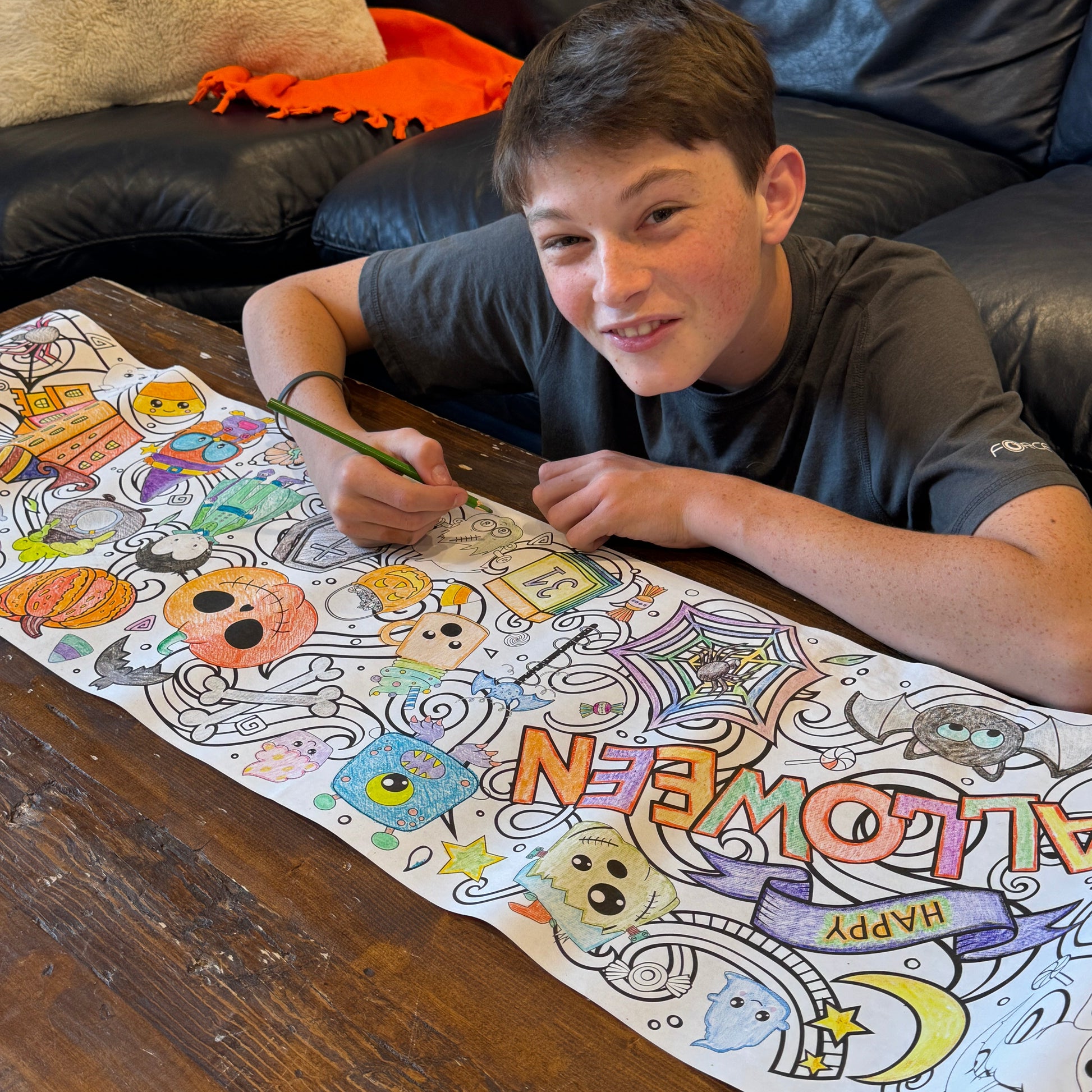 Teen boy coloring a Halloween-themed coloring roll on a wooden coffee table.