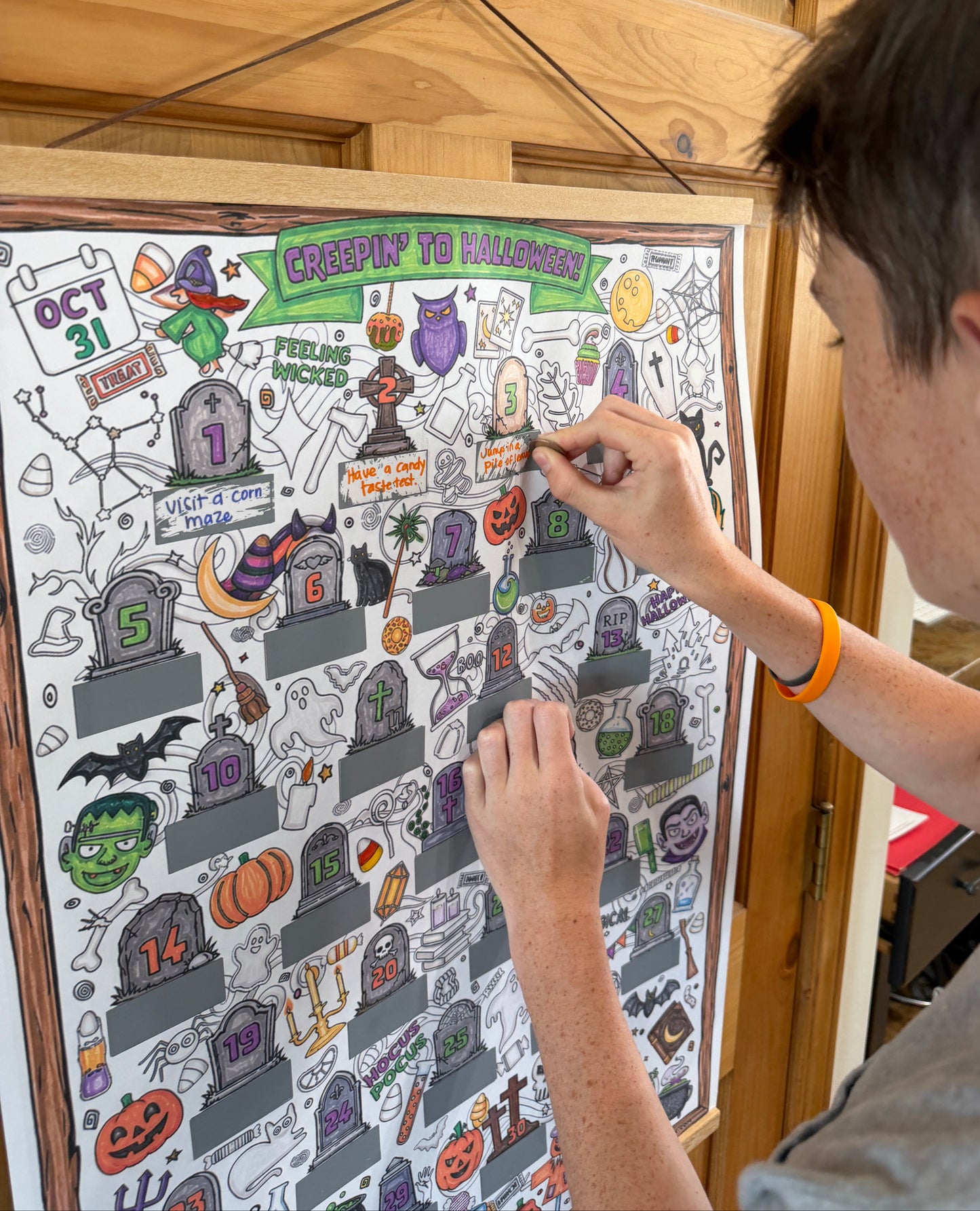 A person coloring in a Halloween-themed countdown or poster with spooky illustrations like tombstones, pumpkins, bats, and ghosts, set against a wooden door background.