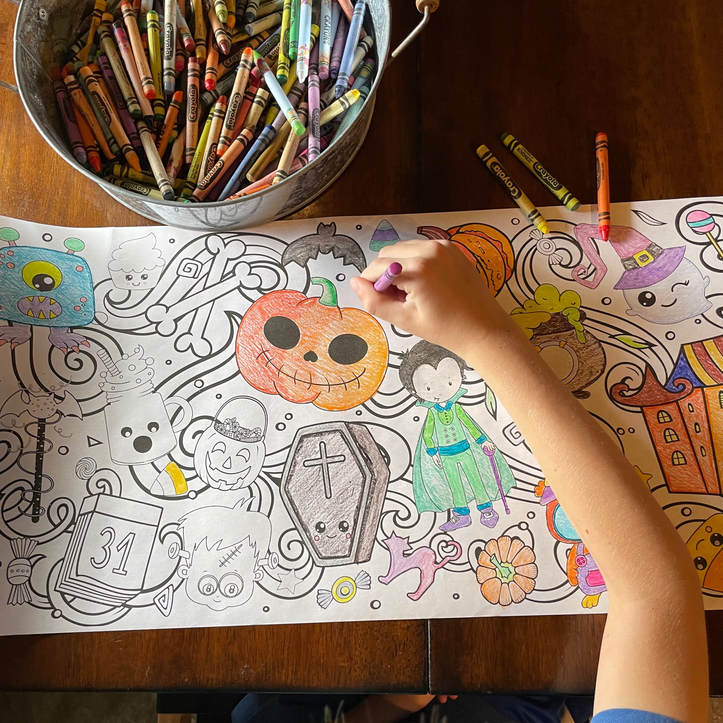 Child coloring a Halloween-themed coloring sheet with crayons on a wooden table.