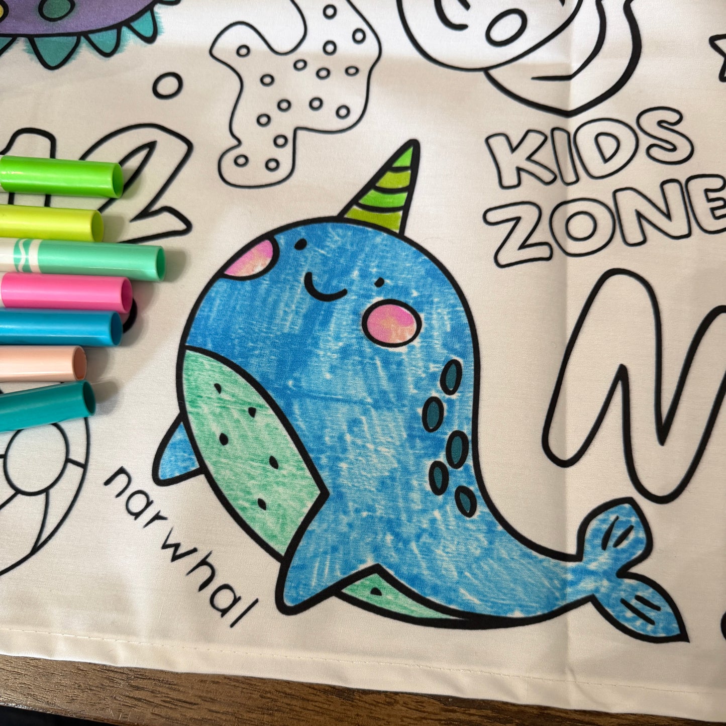 Coloring table cloth with a narwhal design and markers on a wooden table top