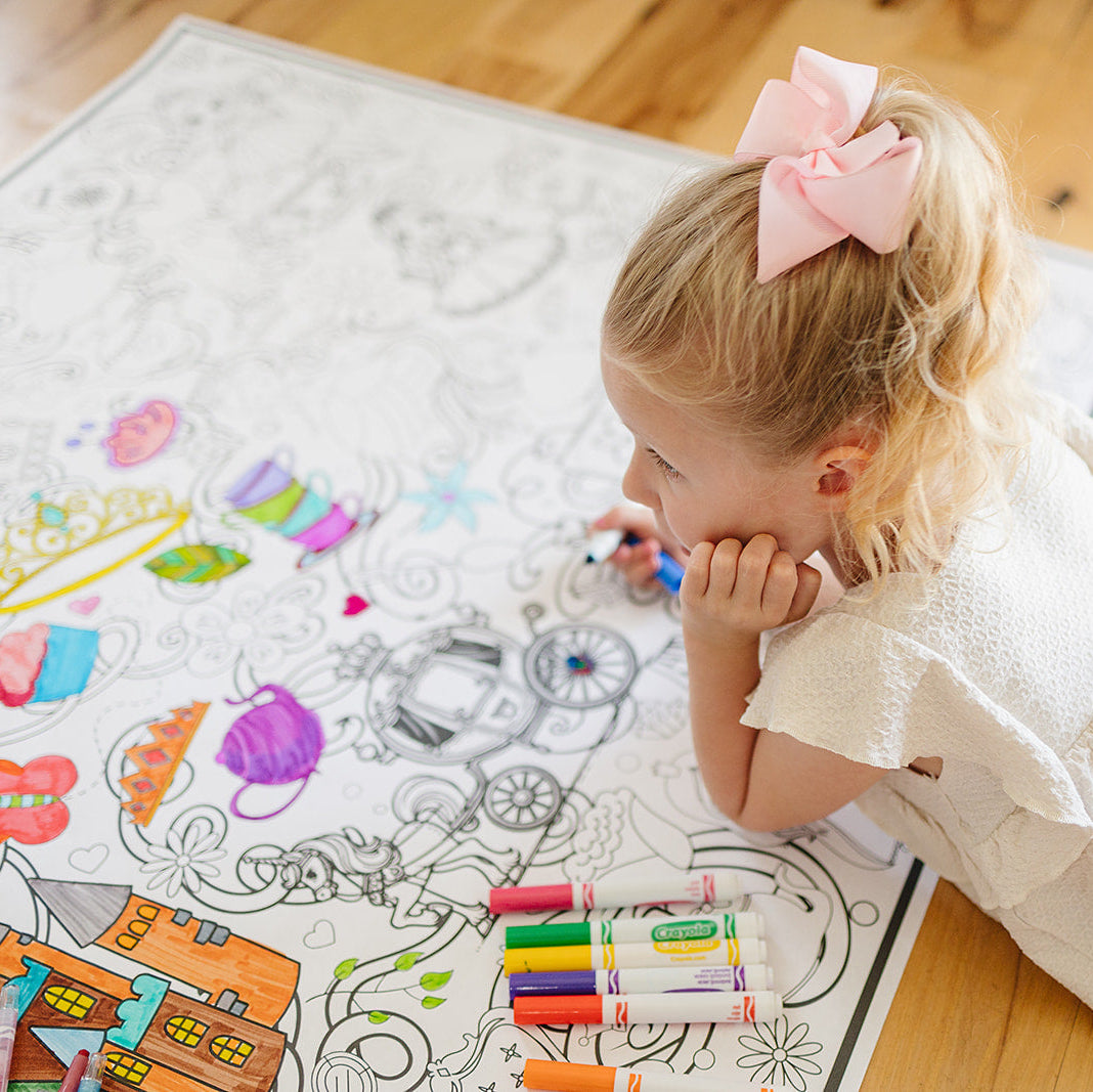 Child coloring on a large princess coloring sheet with crayons on a wooden floor.