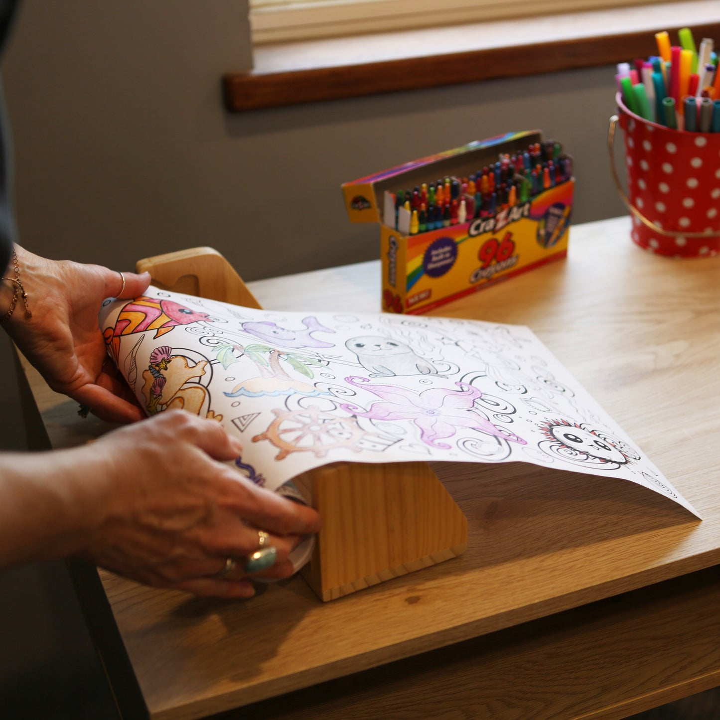 Person putting a coloring roll on a paper dispenser. Paper roll has ocean and underwater designs on it. Theres a box of crayons and bucket of markers on the desk.