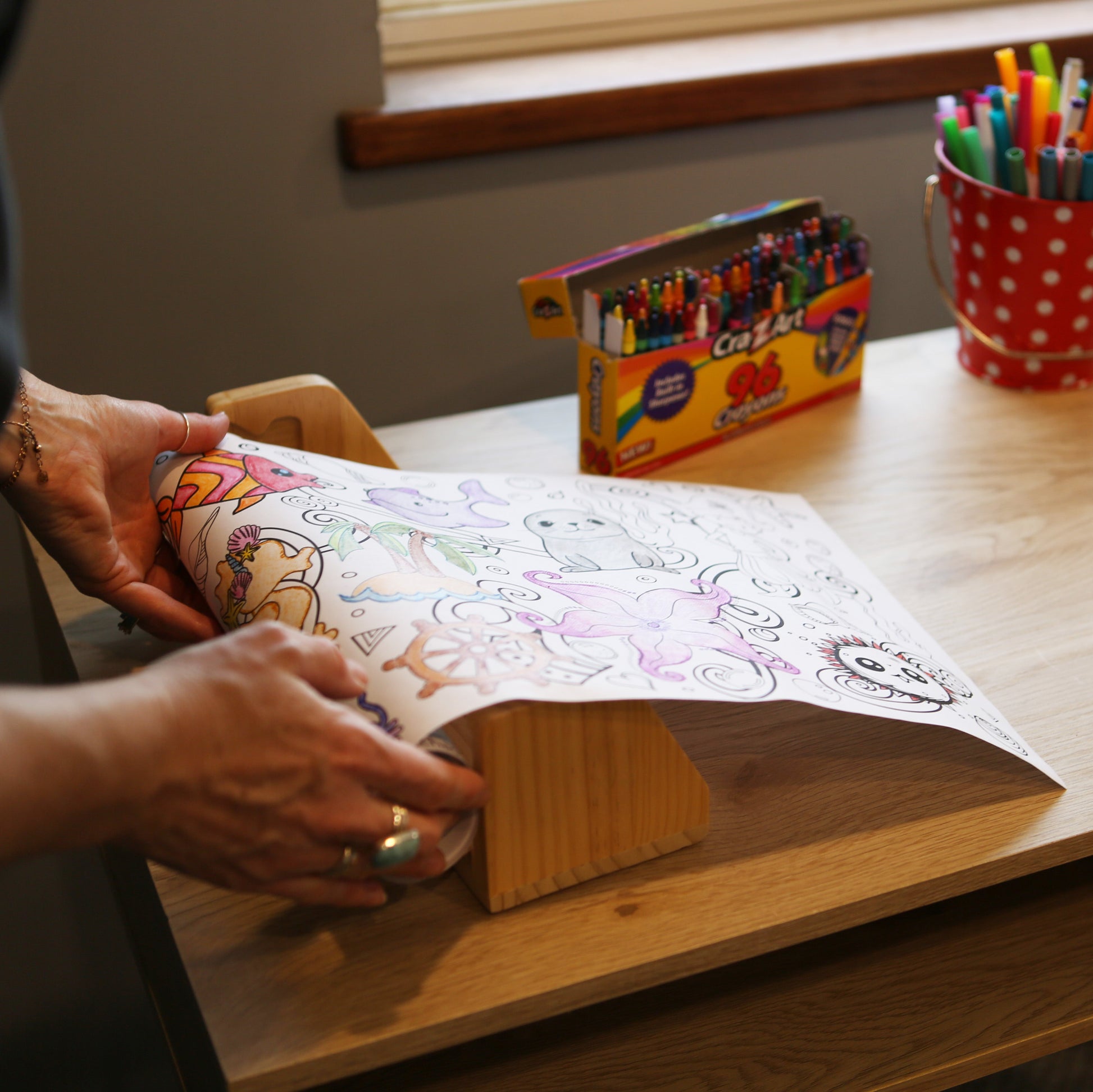 Person putting a coloring roll on a paper dispenser. Paper roll has ocean and underwater designs on it. Theres a box of crayons and bucket of markers on the desk.